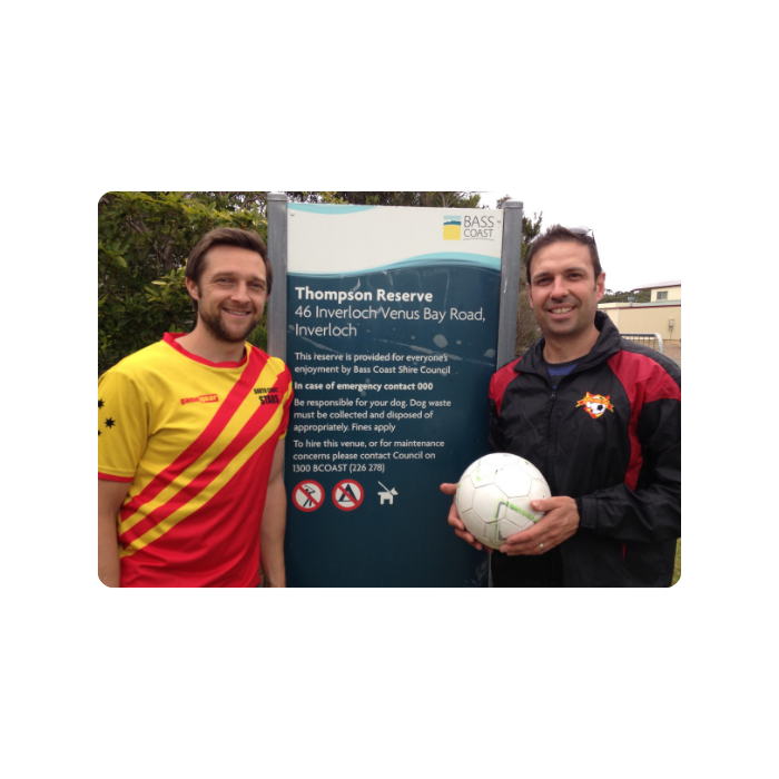 Two men standing outdoors in front of a sign for Thompson Reserve, with one holding a soccer ball. The man on the left is wearing a red and yellow sports jersey, and the man on the right is wearing a black and red jacket.