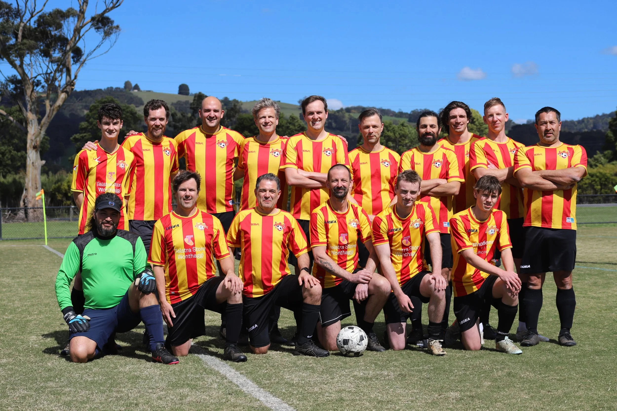 A group of 16 men in soccer uniforms posing for a team photo on a soccer field under a bright blue sky. The team members wear yellow and red striped jerseys, black shorts, and black socks, with one man in a green goalkeeper uniform kneeling in front.
