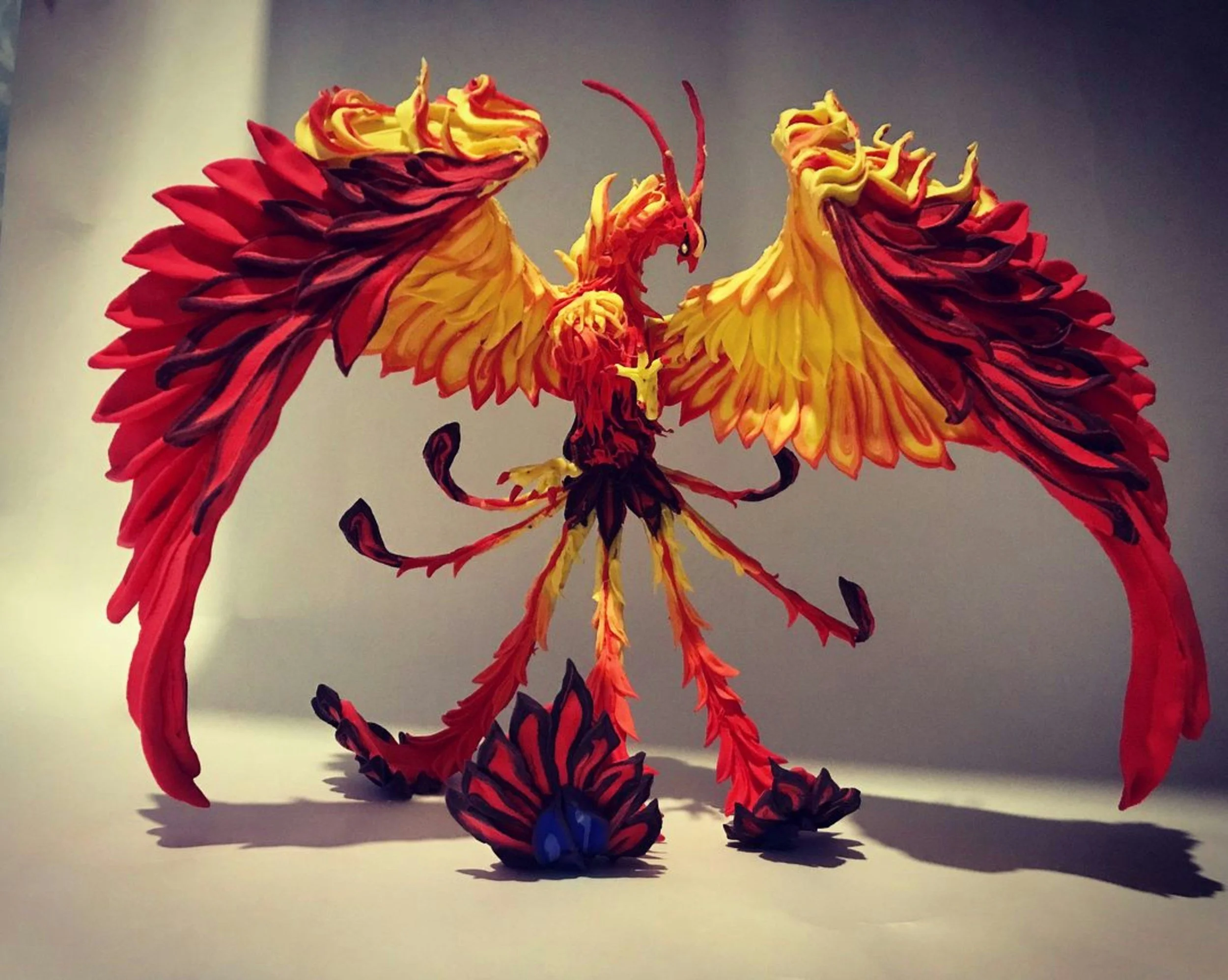Colorful sculpture of a phoenix with outstretched wings, made of layered materials in red, yellow, and black, with detailed feathers and a fierce expression.