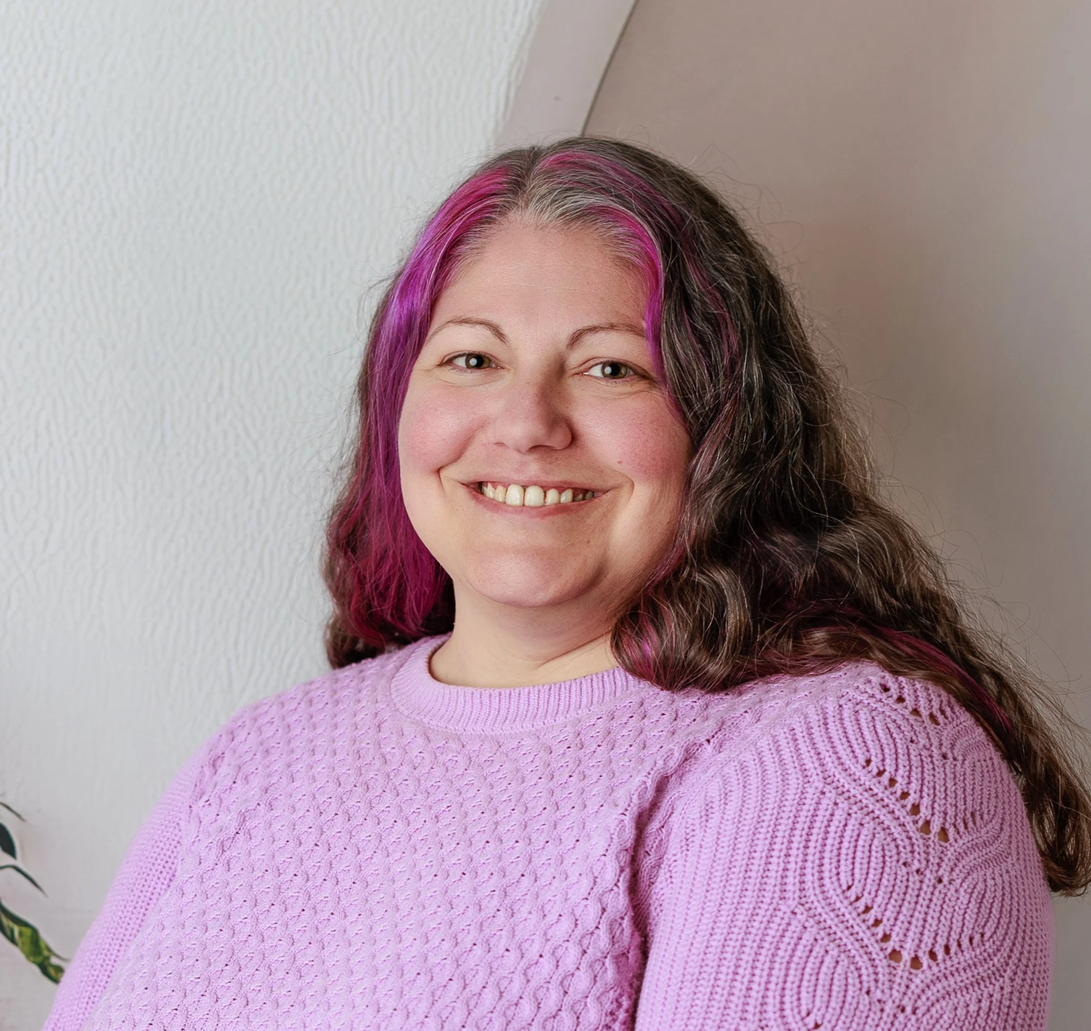 A woman with shoulder-length curly hair, some of which is dyed pink, smiling at the camera, wearing a pink knitted sweater.