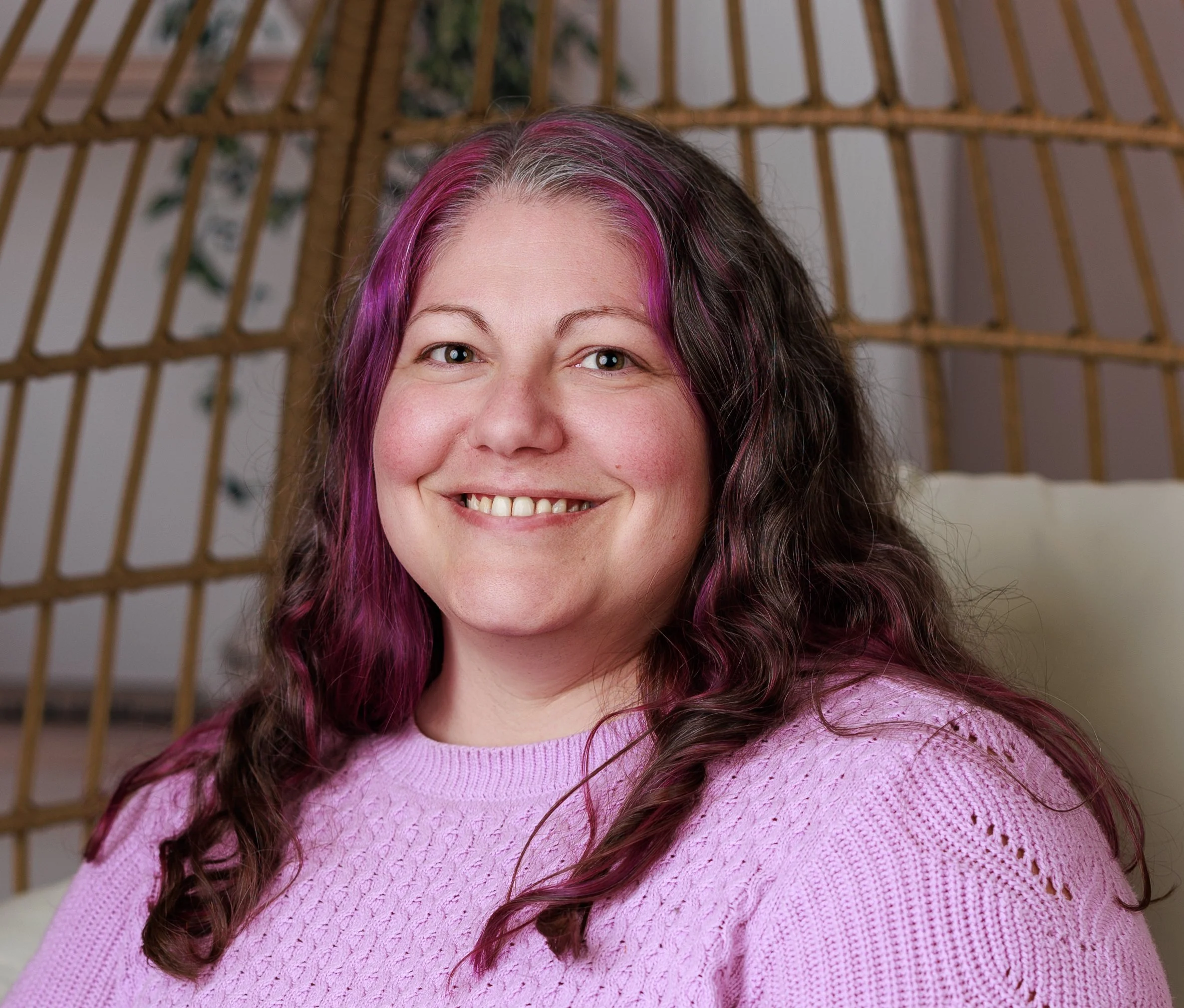 A woman with curly, two-toned hair (pink and dark brown) smiling, wearing a pink knit sweater, sitting against a rattan or wicker background.