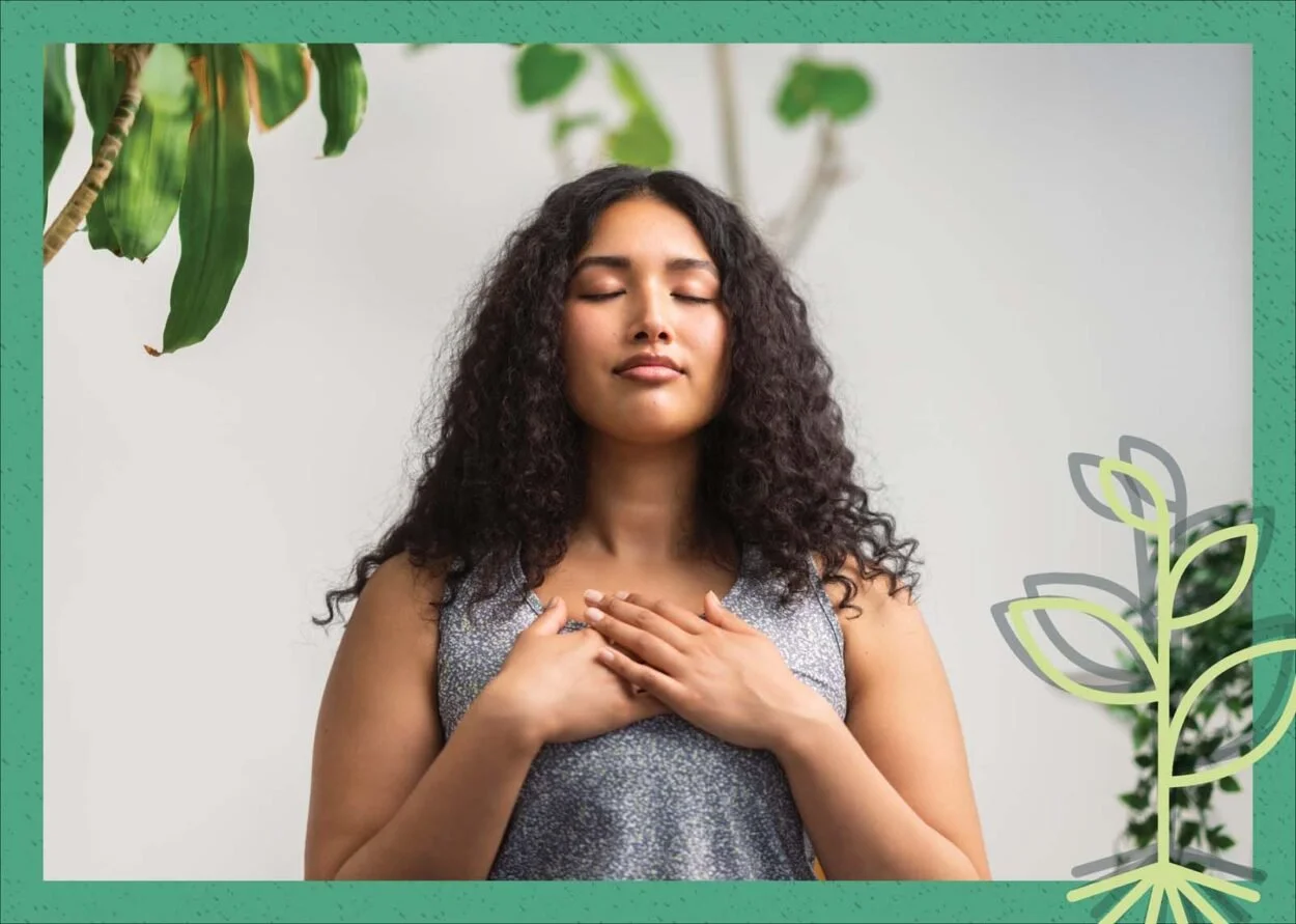 A woman with curly hair meditating with her eyes closed and hands on her chest, surrounded by plants.