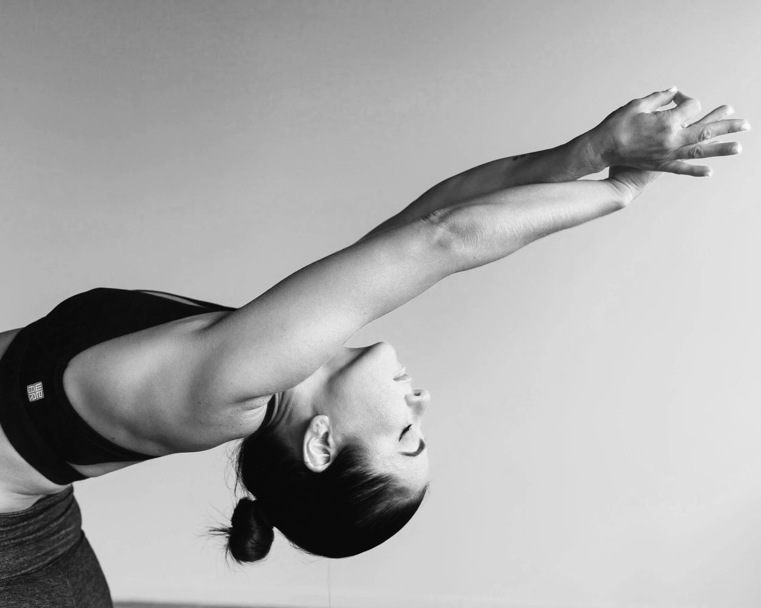A black-and-white photo of a woman practicing yoga, holding a stretch with her head tilted back and arms extended overhead in a pilates and wellness setting.