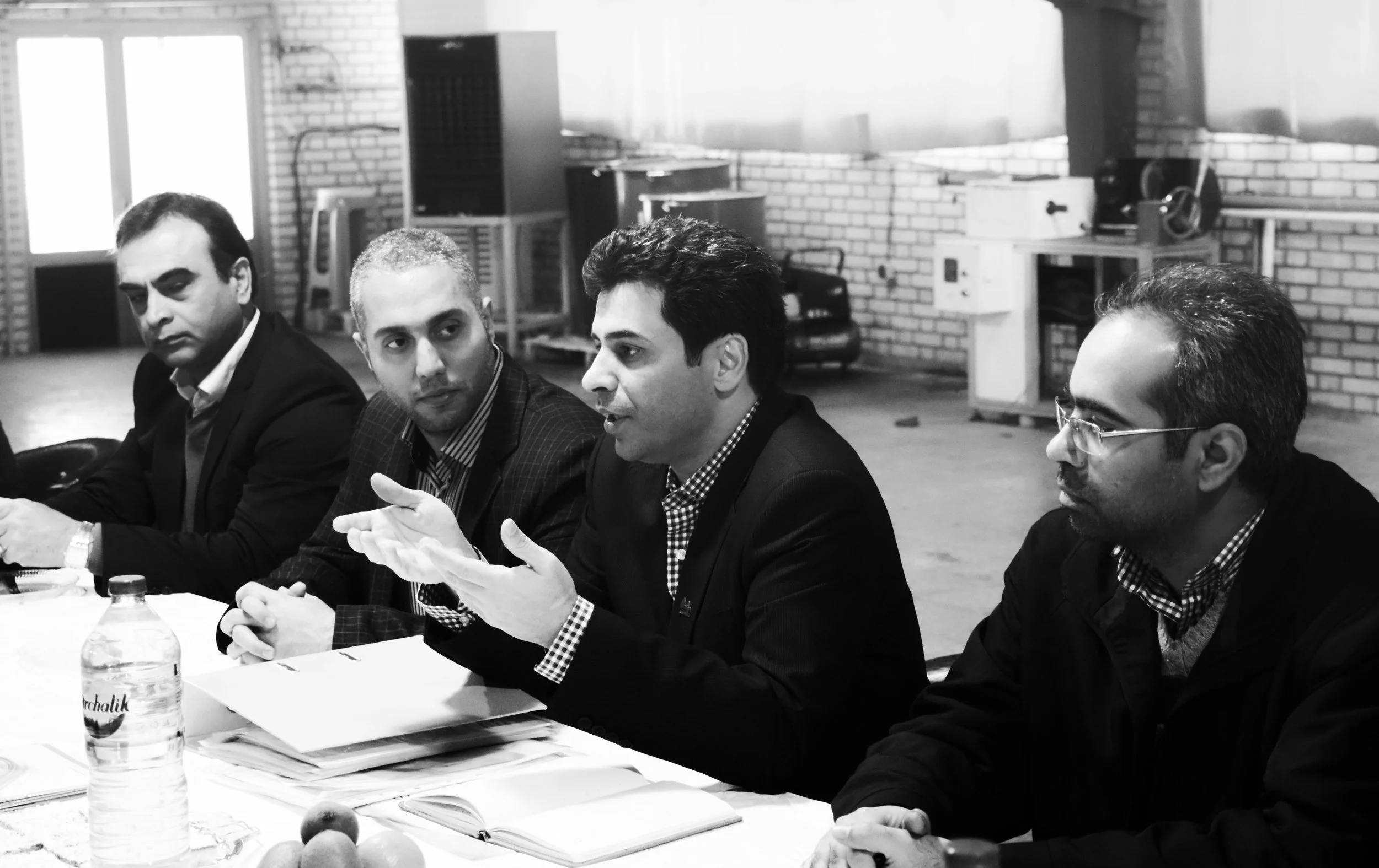 Four men in suits sitting at a meeting table, engaged in discussion, in an industrial-style room with brick walls and large windows.