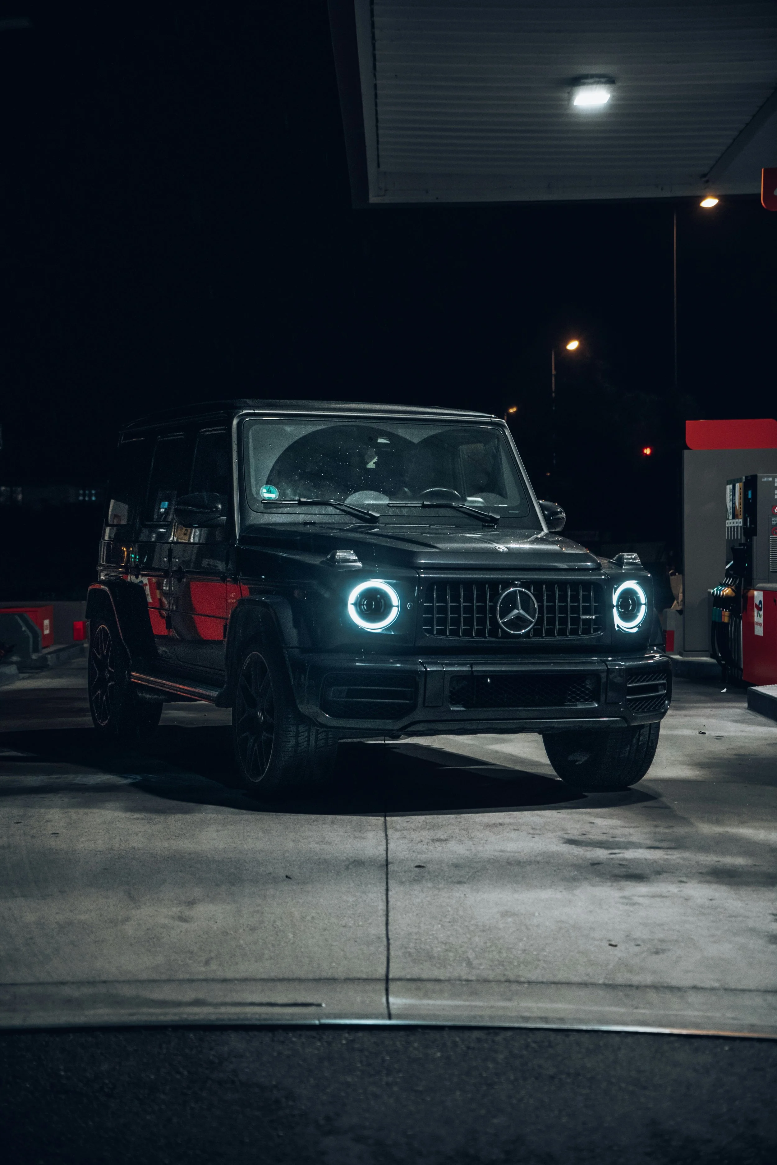 A black Mercedes-Benz G-Class SUV parked at a gas station at night, with illuminated headlights and surrounding fuel pumps.