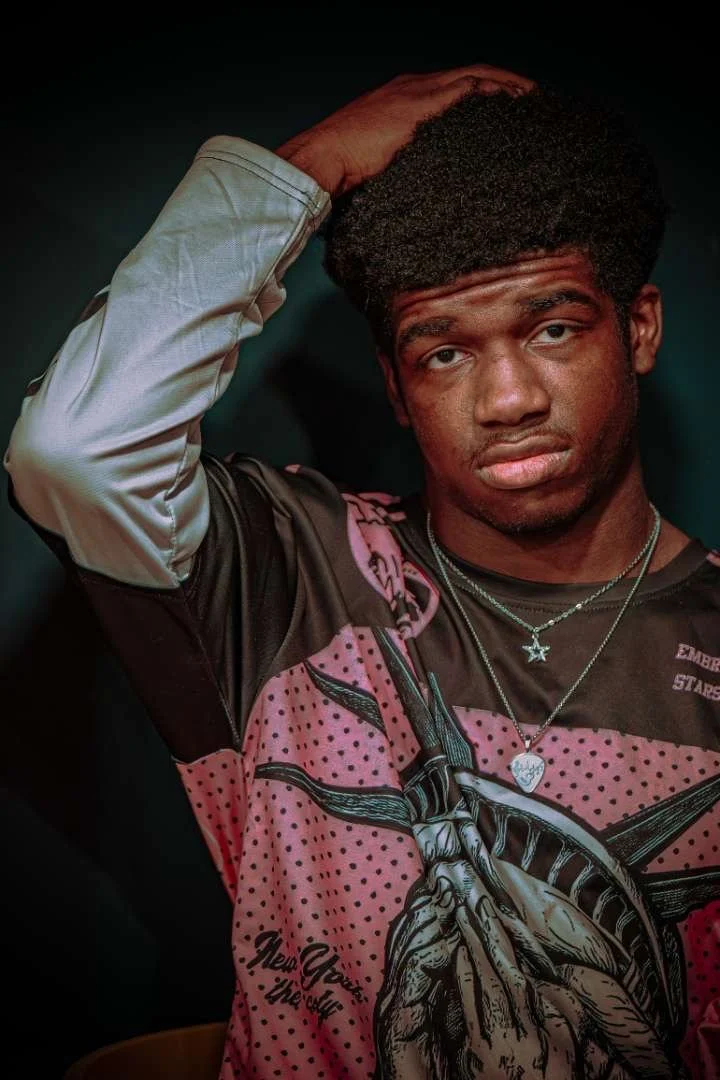 Young man with dark curly hair, holding his head with one hand, wearing a black and pink graphic shirt with a large hand illustration, multiple necklaces, and a serious expression, against a dark background.