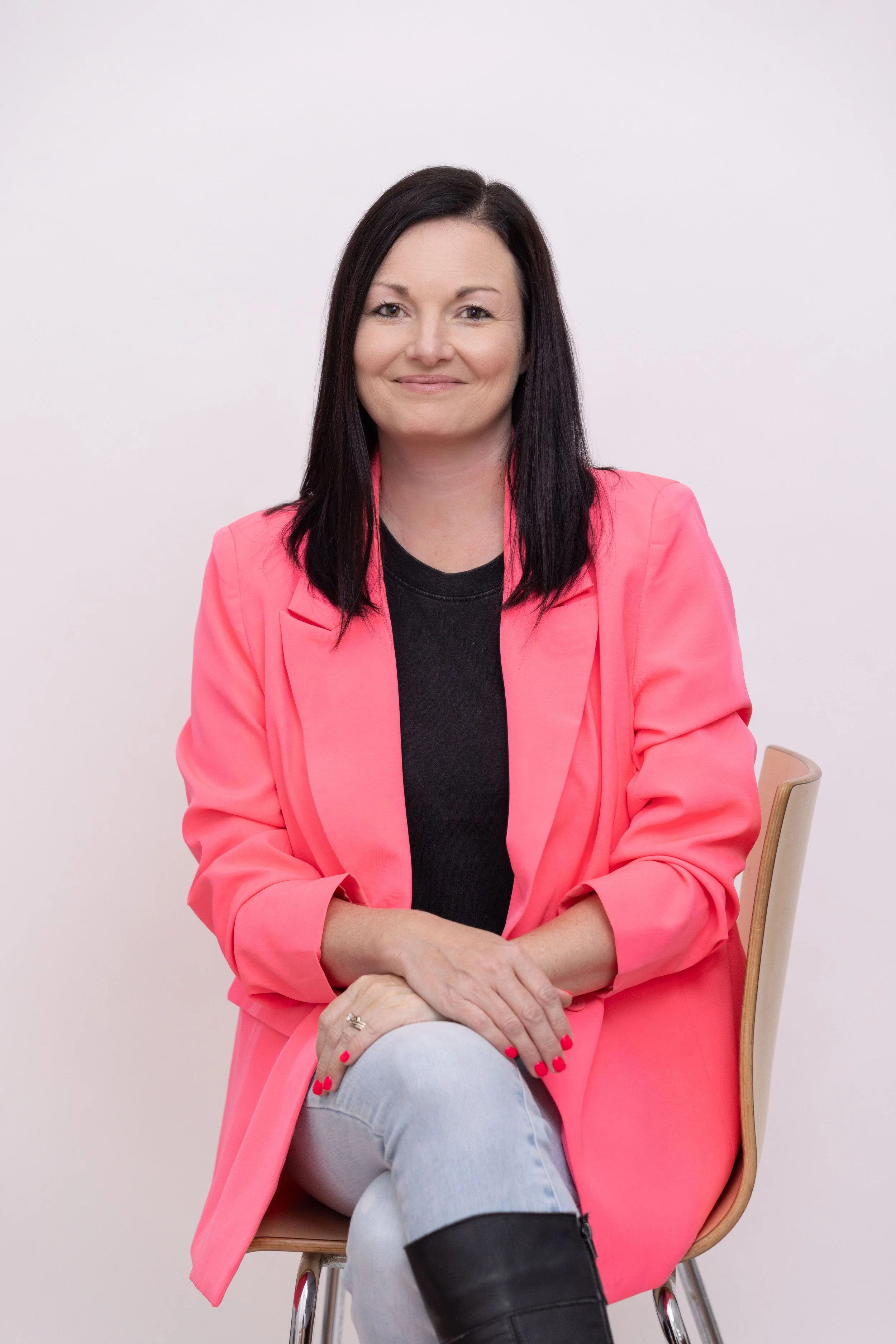 A woman with straight dark hair and fair skin sitting on a wooden chair against a plain white background. She is wearing a bright pink blazer over a black top, gray jeans, and a black boot on her left leg. She is smiling gently, with her right hand resting on her knee and her left hand on her lap.
