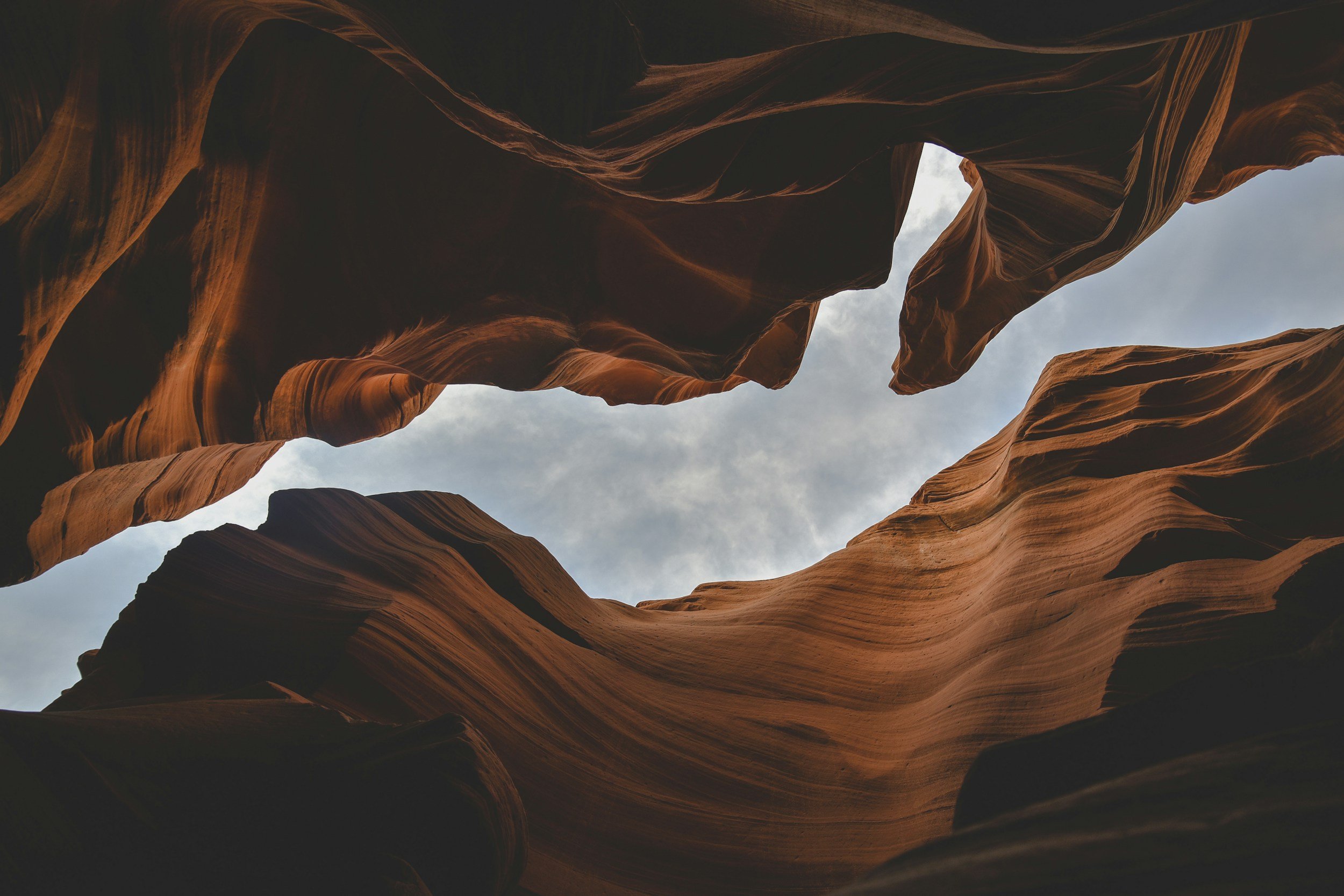 View of a slot canyon with tall, undulating reddish-brown rock walls and a cloudy sky above.