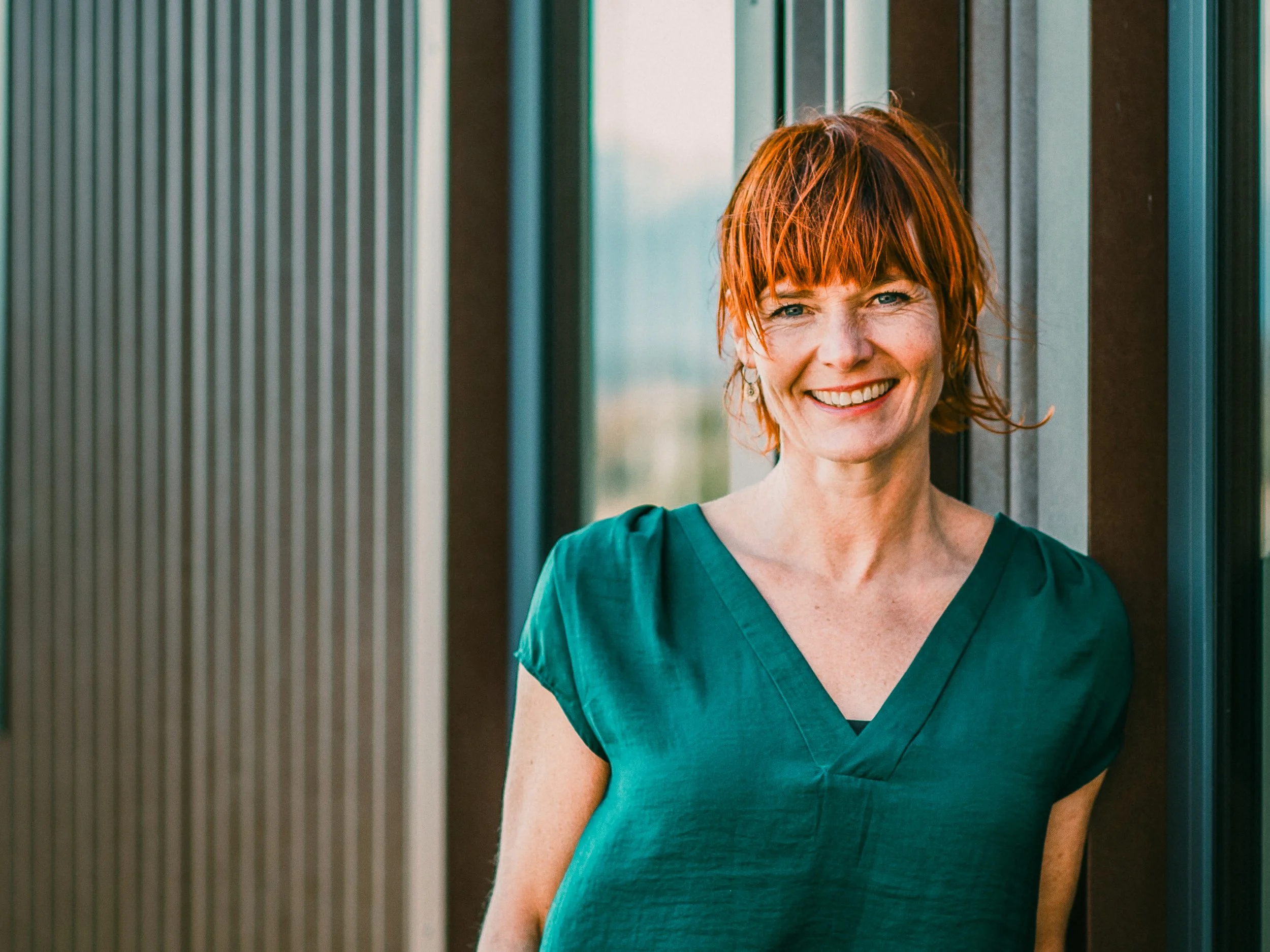 A woman with short red hair, wearing a green top, smiling while standing outdoors beside a modern building with glass windows.