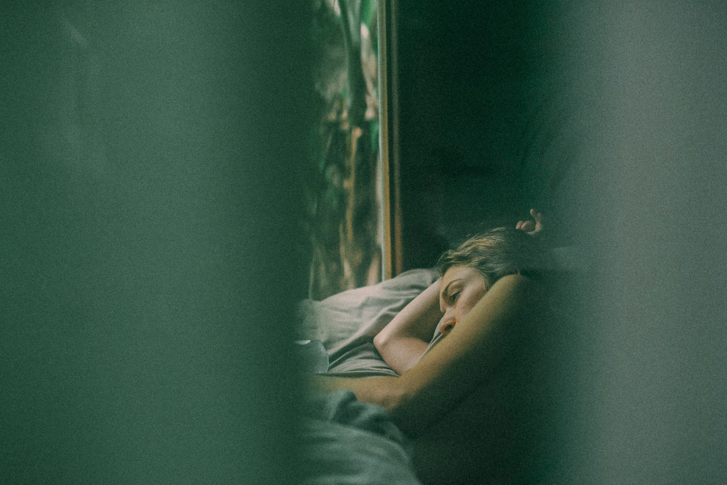 A young woman lying in bed, resting her head on a pillow, with her eyes open, in a dimly lit room, viewed through a slightly blurred or obscured perspective.