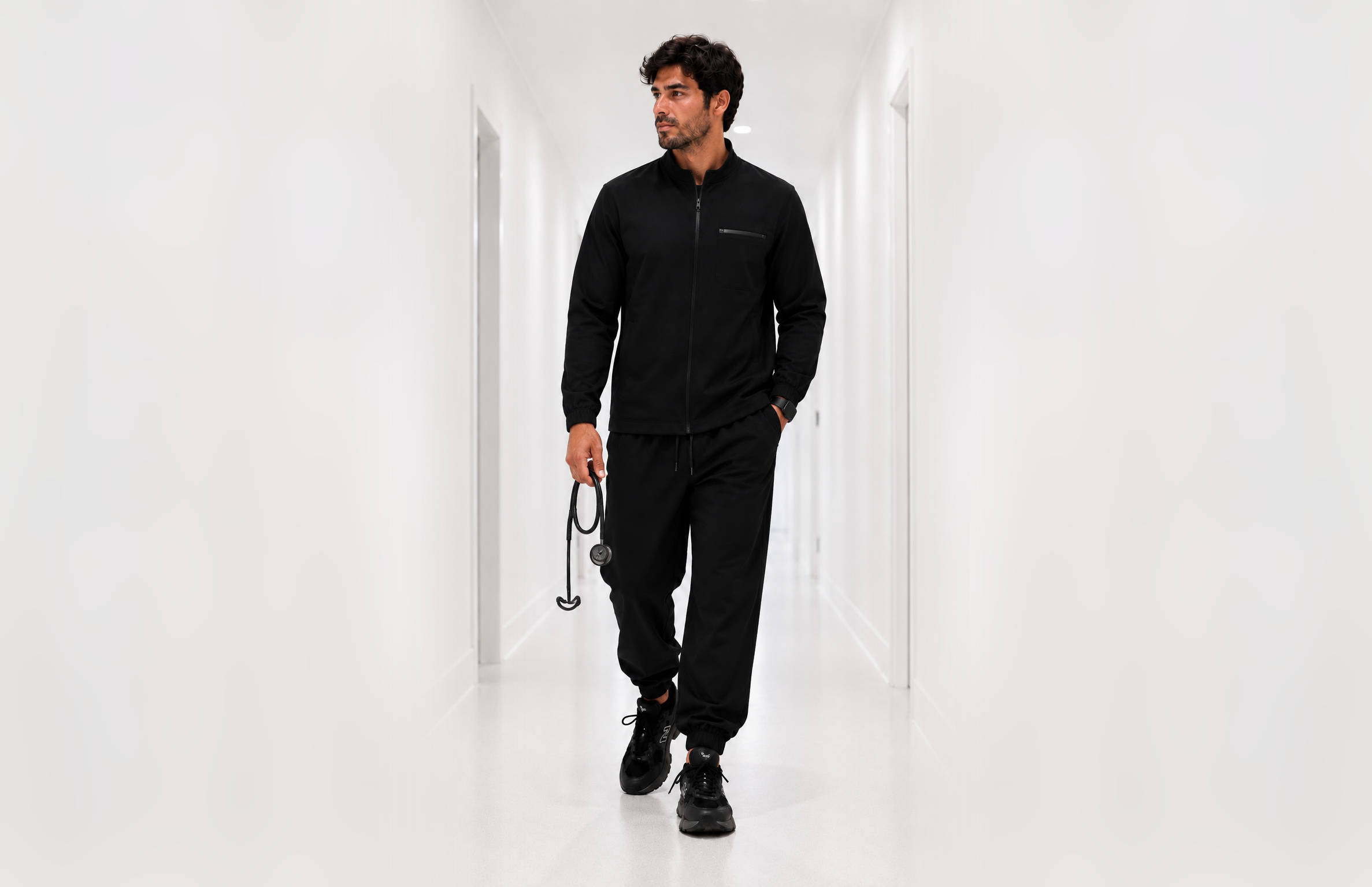 A man walking down a white hallway holding a stethoscope in his right hand, wearing a black bomber jacket, black pants, and black sneakers.