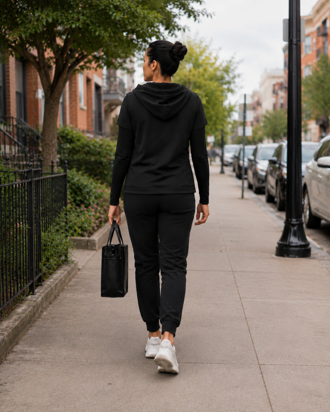 A woman dressed in black jogging pants, a black hoodie, and white sneakers, walking down a city sidewalk while carrying a black tote bag.