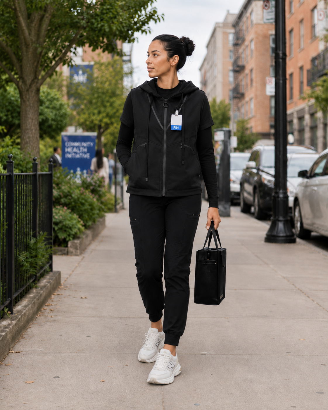 A woman walking on a city sidewalk with an RN badge, carrying a black bag, dressed in black athletic clothes and sneakers, with greenery and buildings in the background.