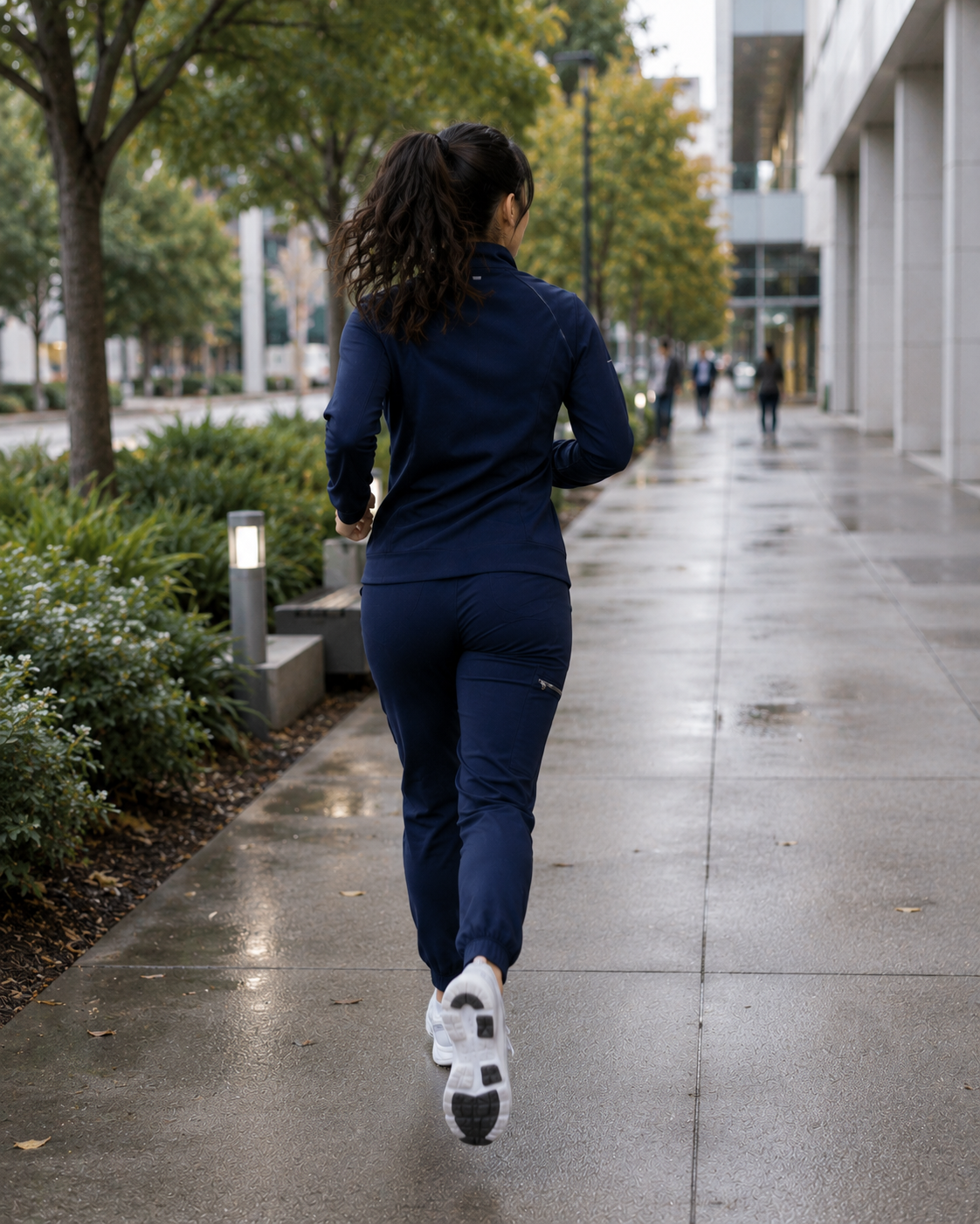 A woman jogging on a city sidewalk during overcast weather, wearing a blue athletic outfit and white running shoes, with trees and buildings along the street.