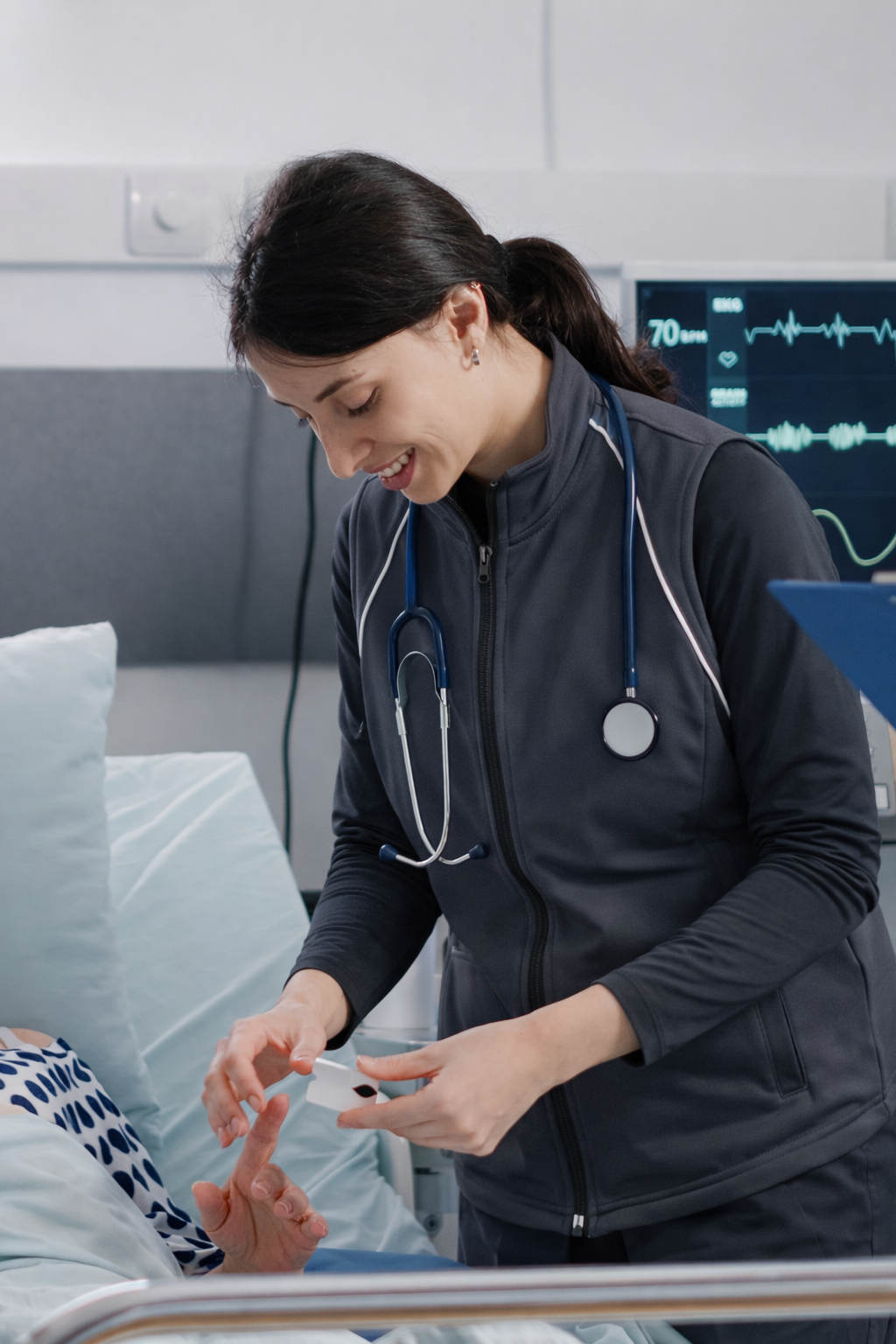 A female nurse in gray scrubs and a stethoscope placing a wristband on a patient's hand in a hospital room.