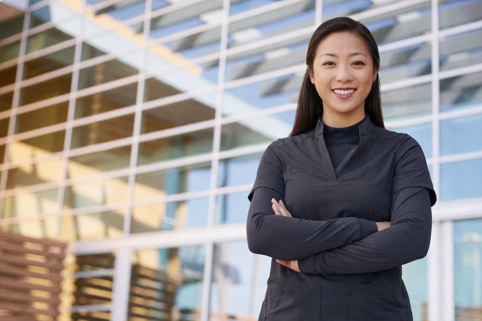A confident woman with dark hair, wearing a black athletic jacket, standing with arms crossed in front of a modern glass building.