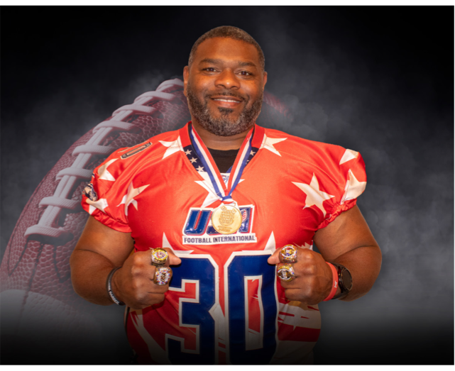 Smiling man in a red American football jersey with white stars, wearing a gold medal. He is making fists with both hands, wearing rings, and standing in front of a football background.