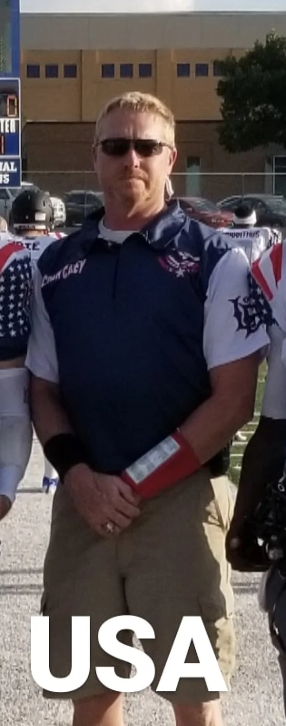 A man standing outdoors in front of a building, wearing sunglasses, a navy sleeveless vest with American flag themed sleeves, and khaki shorts, with the text 'USA' at the bottom.