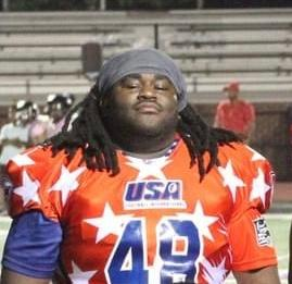 A football player in a red, white, and blue uniform with stars, wearing a grey head covering, standing on a football field.