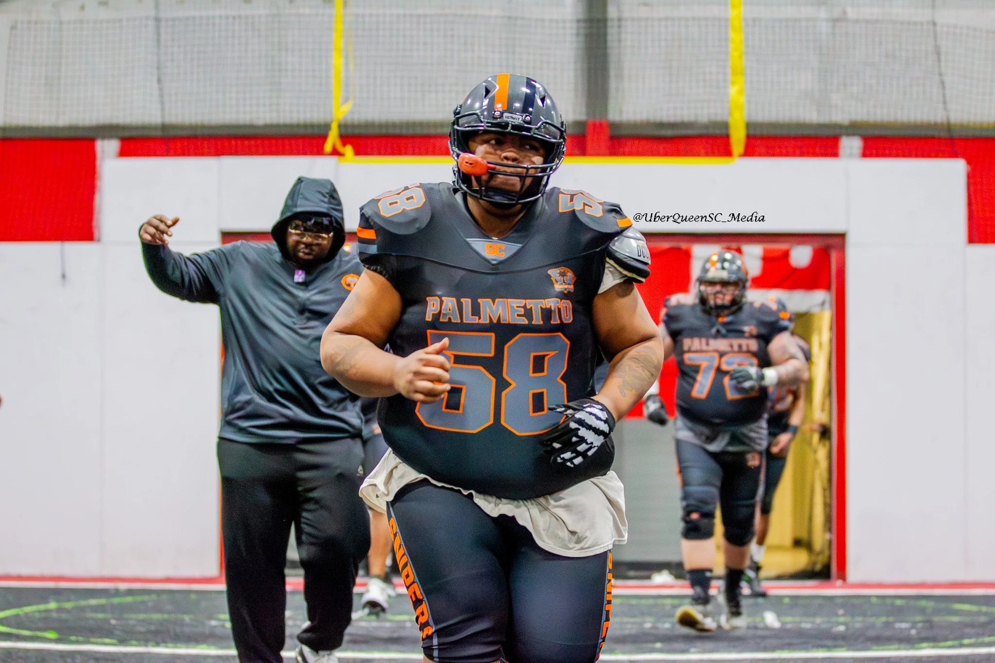 Football players from Palmetto team entering the field through an indoor tunnel, wearing black and orange uniforms with helmets.