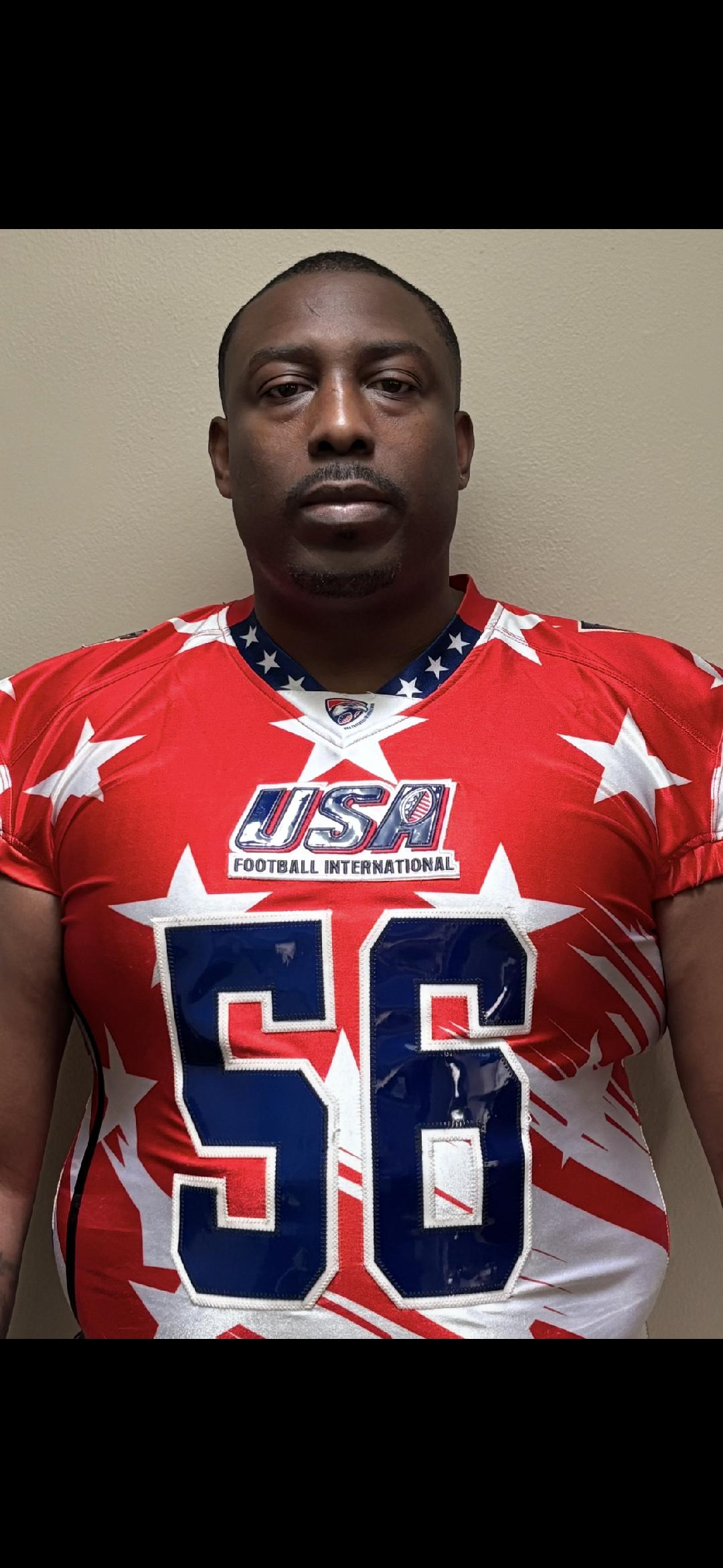 A man in a red USA-themed football jersey with white stars and blue number 56, standing against a beige wall.