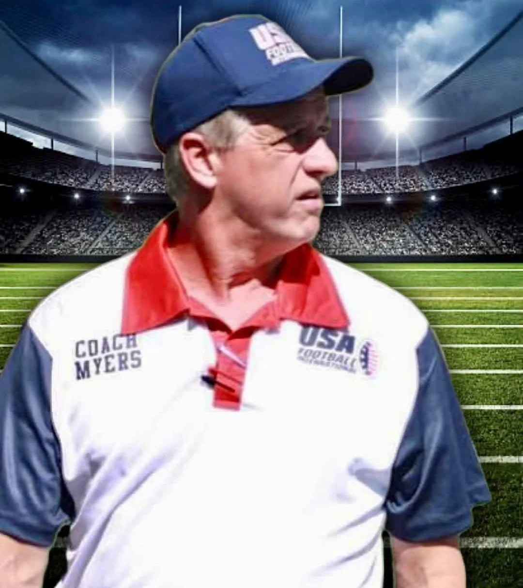 A man in a USA football coach uniform looking to the right on a football field with a stadium in the background under cloudy skies.