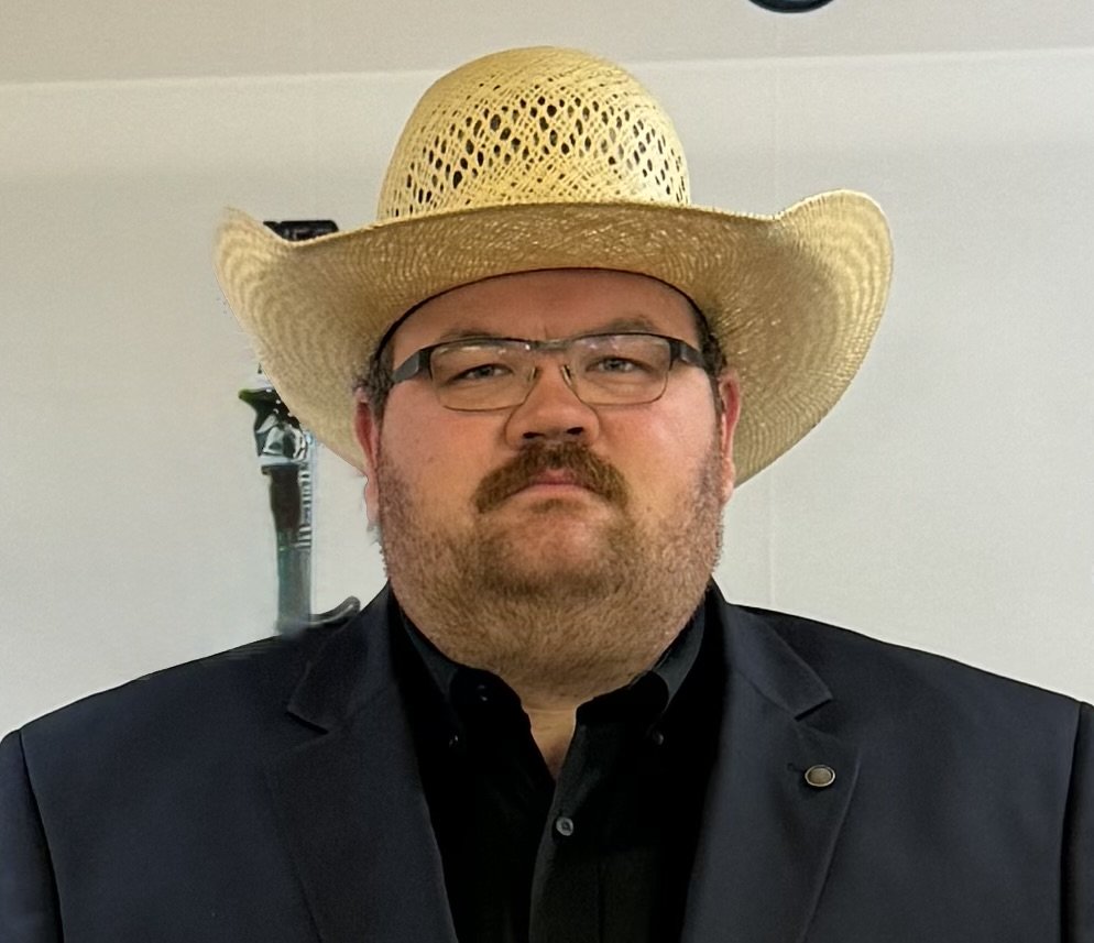 A man wearing a black suit with a bolo tie, glasses, and a wide-brimmed straw hat, standing indoors against a plain light-colored wall.