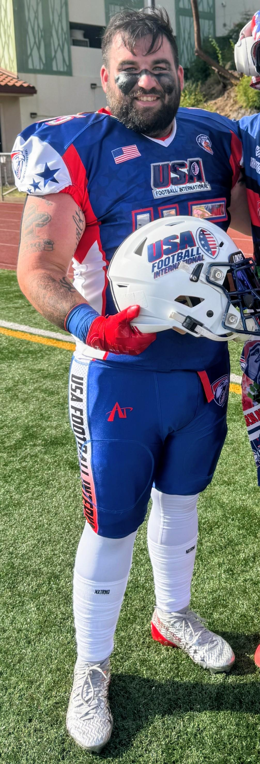A smiling American football player in blue, red, and white uniform holding a white helmet with 'USA Football International' logo, standing on a football field.