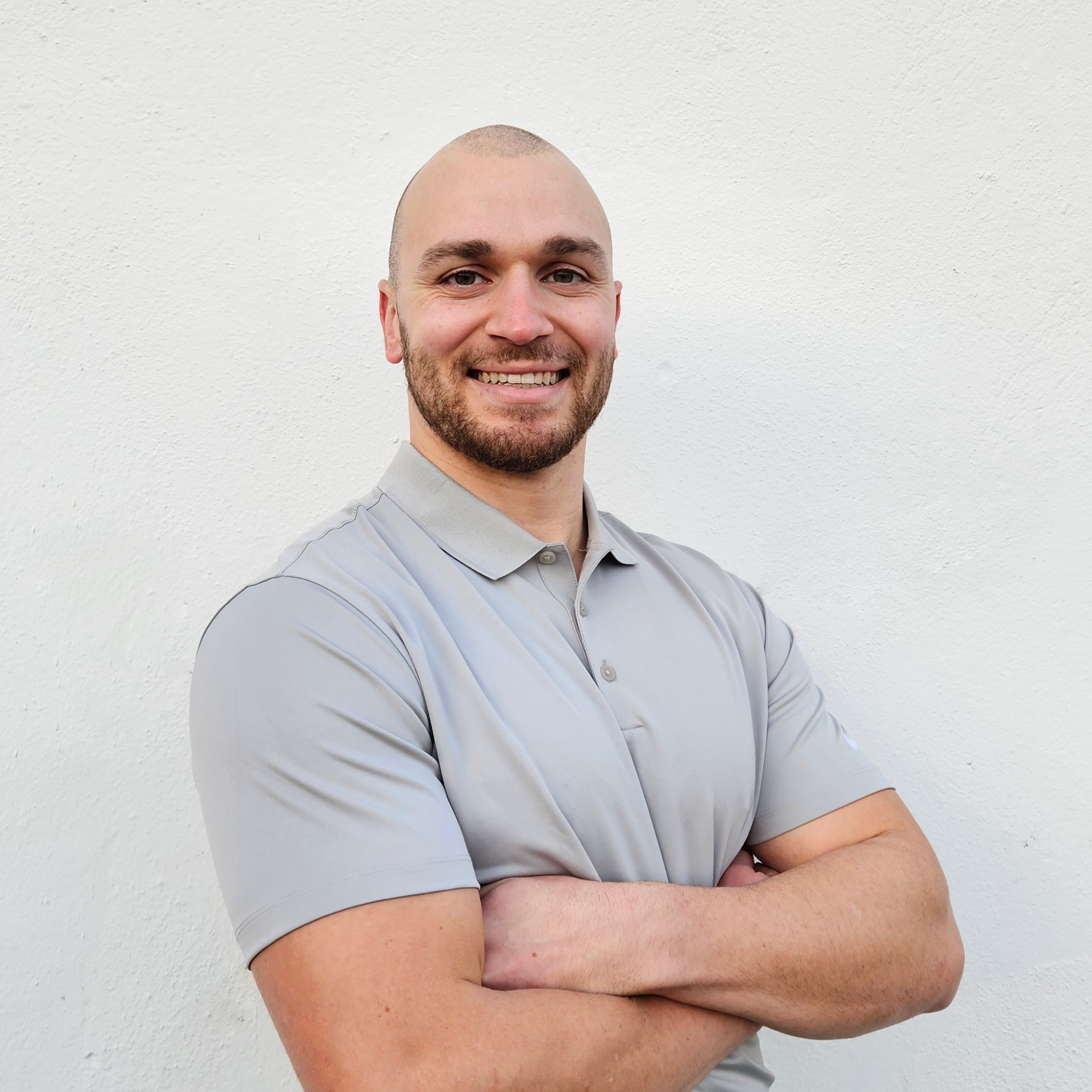 A smiling man with a beard and a shaved head, wearing a light gray polo shirt, standing with arms crossed in front of a plain white wall.