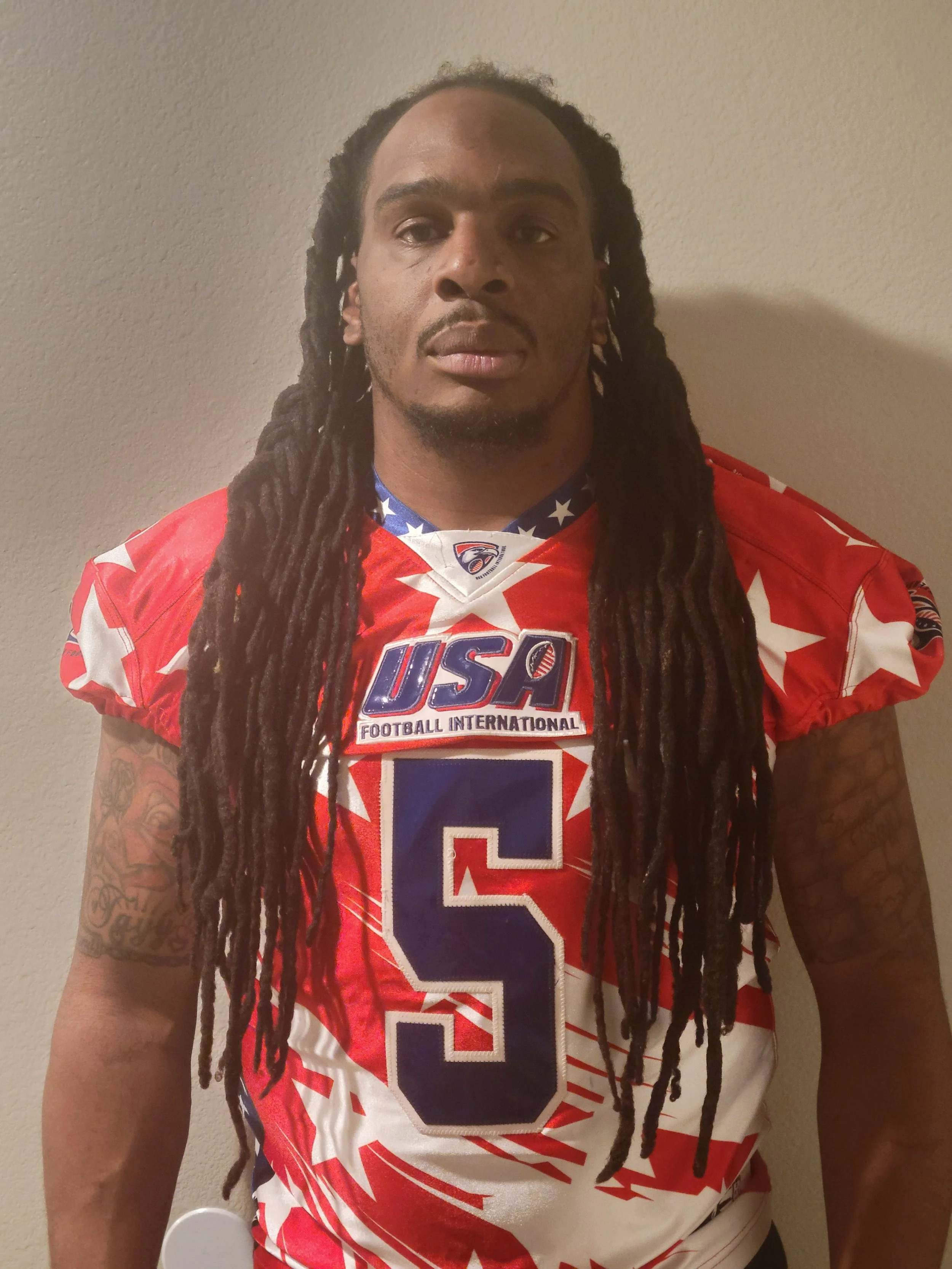 A man in a red, white, and blue USA football jersey with the number 5, standing against a plain wall.