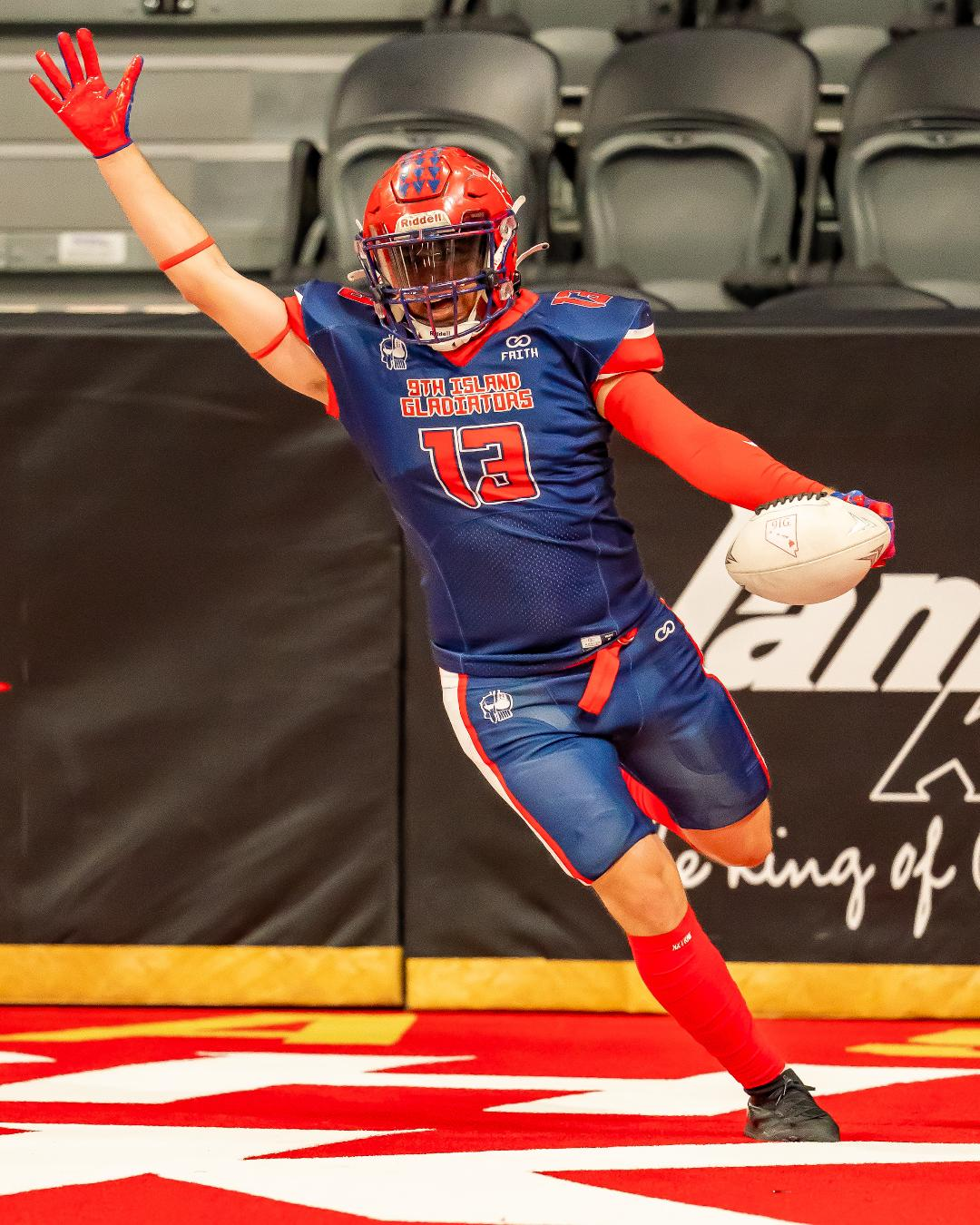A football player celebrating after scoring, wearing a blue and red uniform with the text '9th Island Gladiators' and jersey number 13, holding a football in one hand, on a football field.