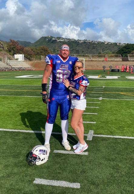 A football player in a blue USA-themed uniform standing on a football field next to a woman in casual white shorts and a USA-themed shirt. They are posing together with the football player's arm around her. The football helmet is on the ground in front of him, and the background shows bleachers, green hills, and a partly cloudy sky.