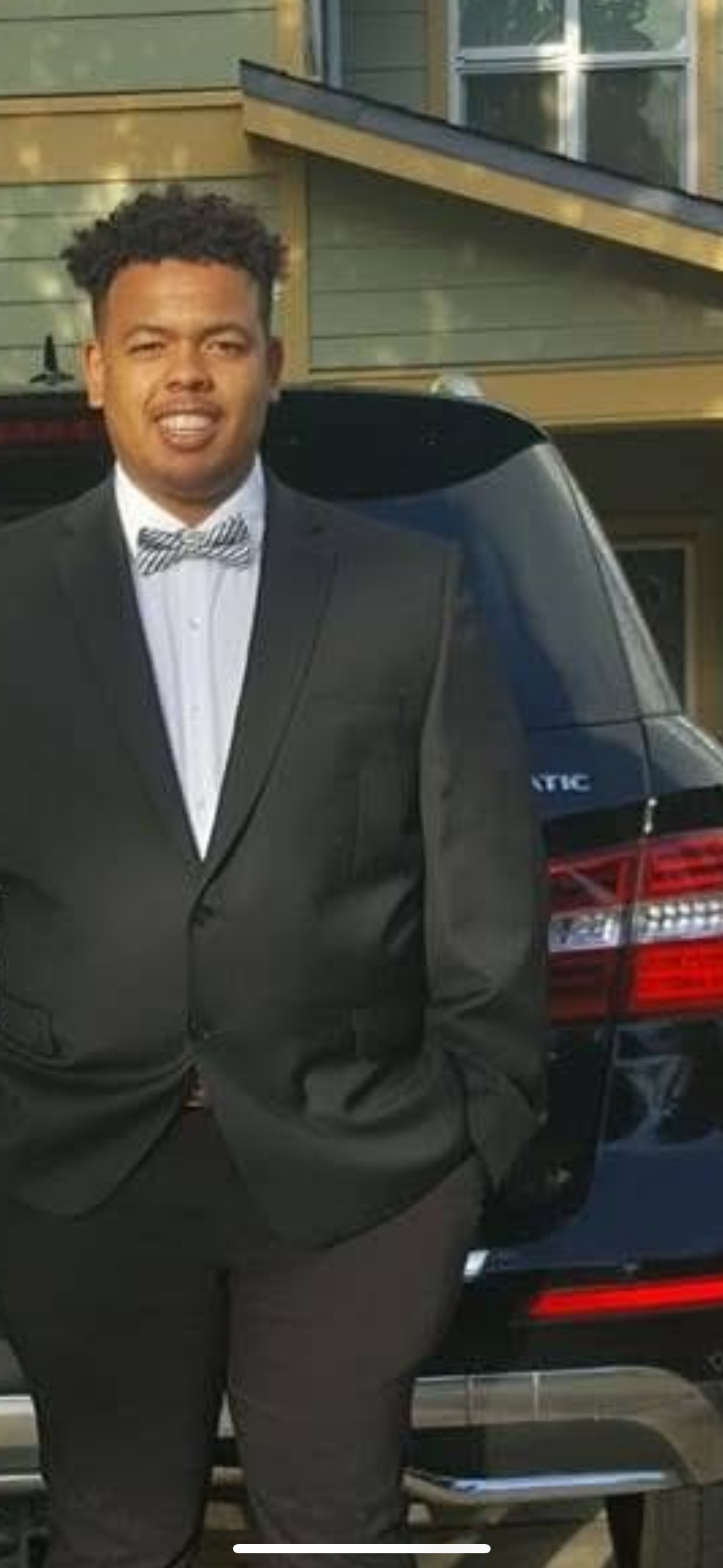 A young man dressed in a black tuxedo with a bow tie, standing outdoors next to a black vehicle, with a house in the background.