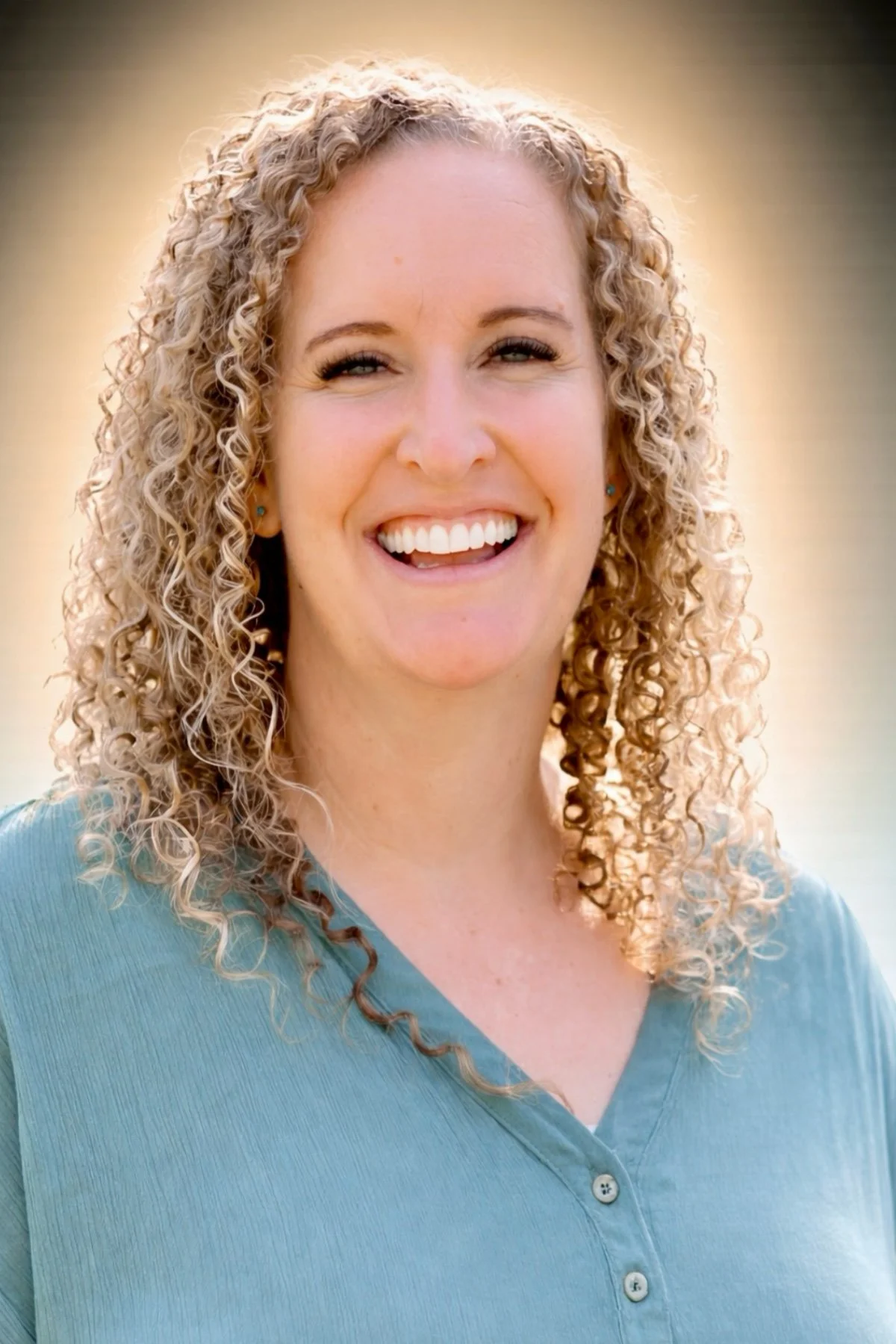A woman with curly blonde hair smiling outdoors in a light blue top.
