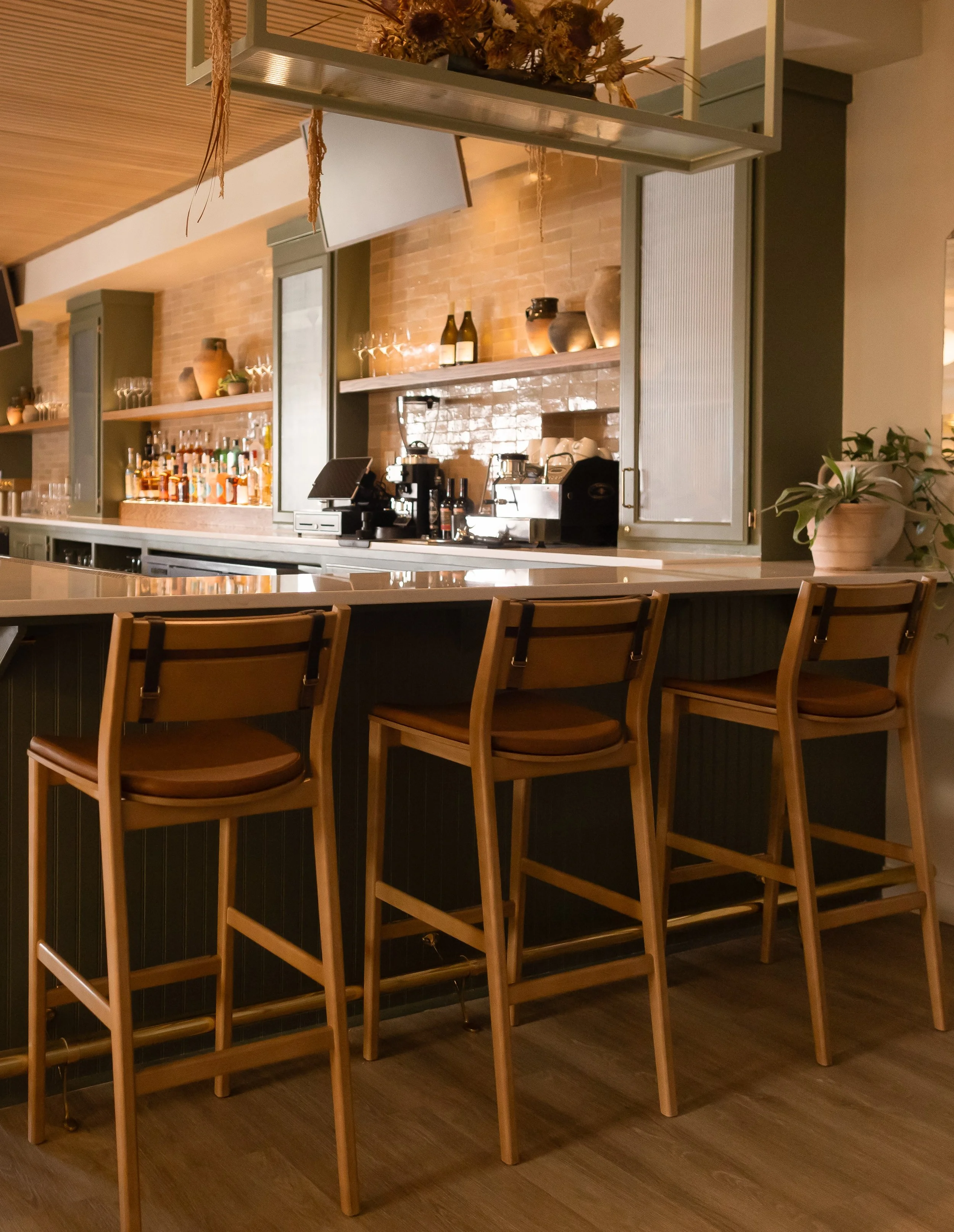 Bar area with wooden bar stools, shelves with bottles and glasses, coffee machines, and decorative pottery against a brick wall.