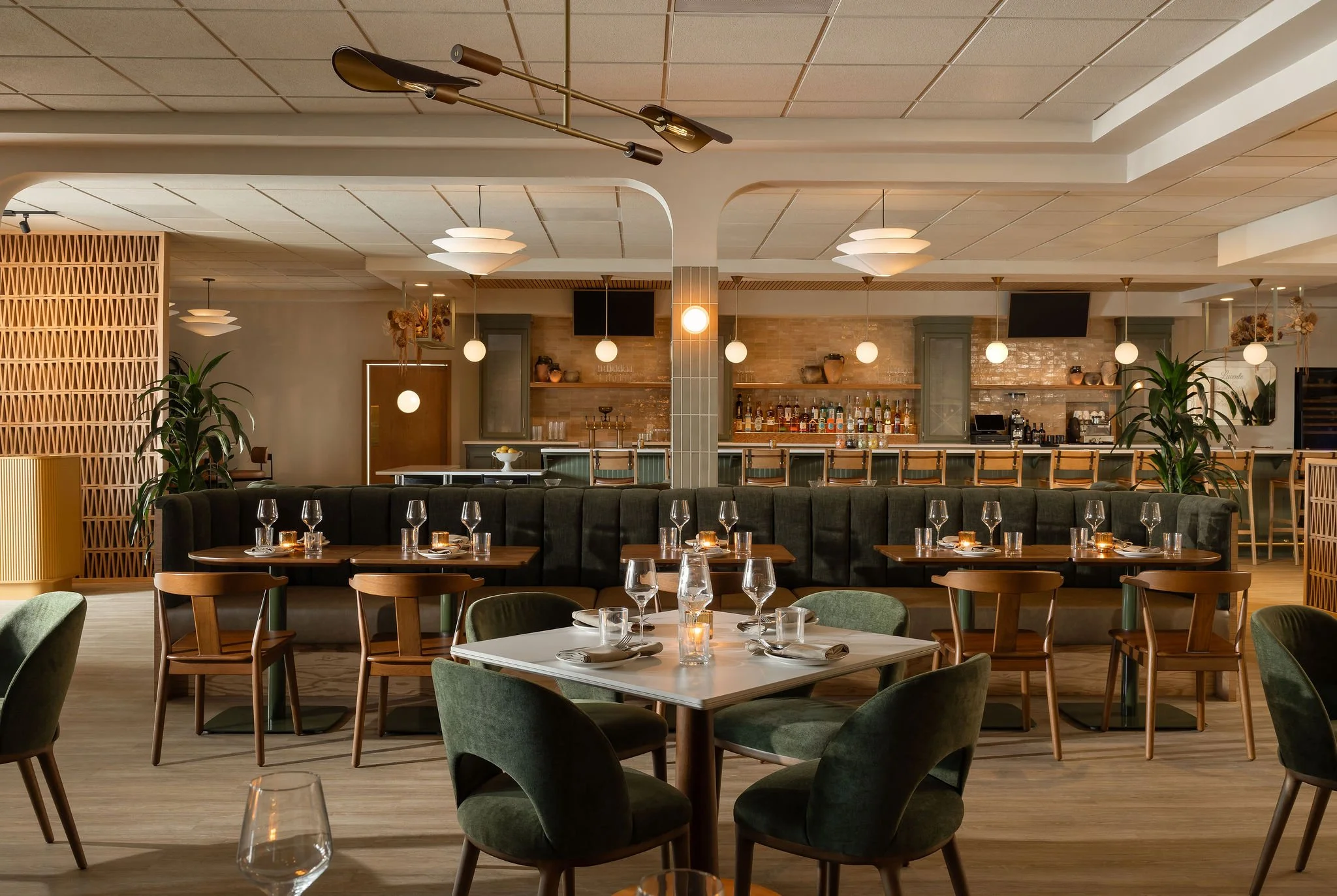 Elegant restaurant interior featuring tables with green chairs, candles, wine glasses, a bar in the background with bottles, potted plants, and modern ceiling lights.