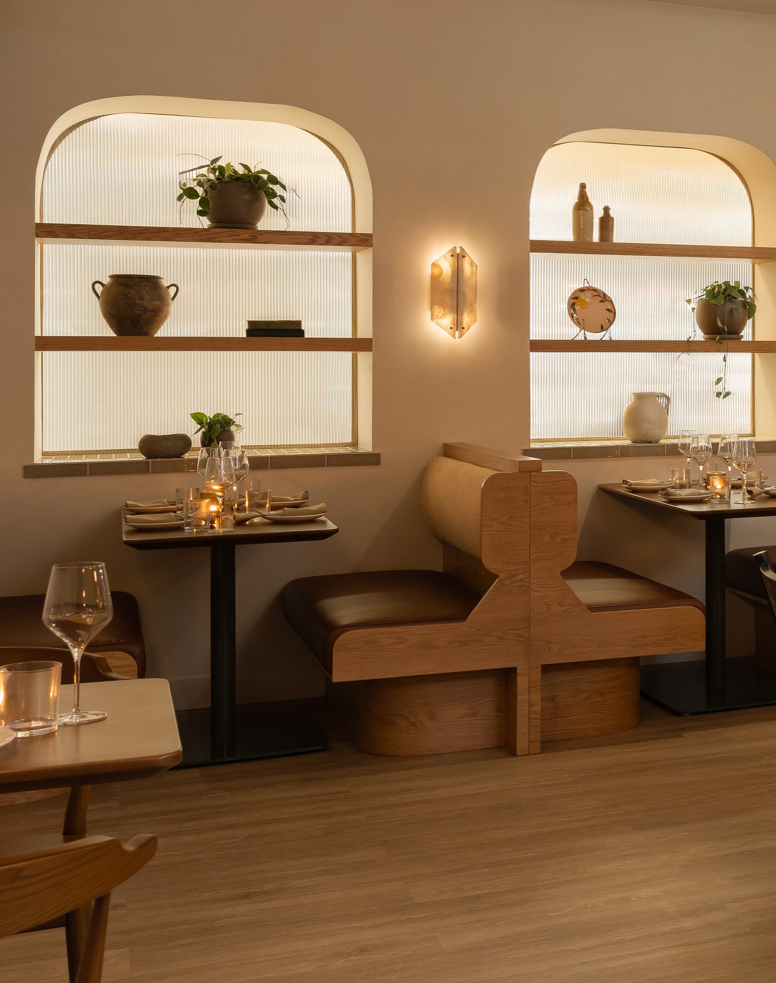Interior of a restaurant or cafe with two arched wall niches illuminated with backlighting. The left niche has a potted plant with green leaves, a clay pot, and a small stack of black books. The right niche has two bottles, a decorative plate with ye