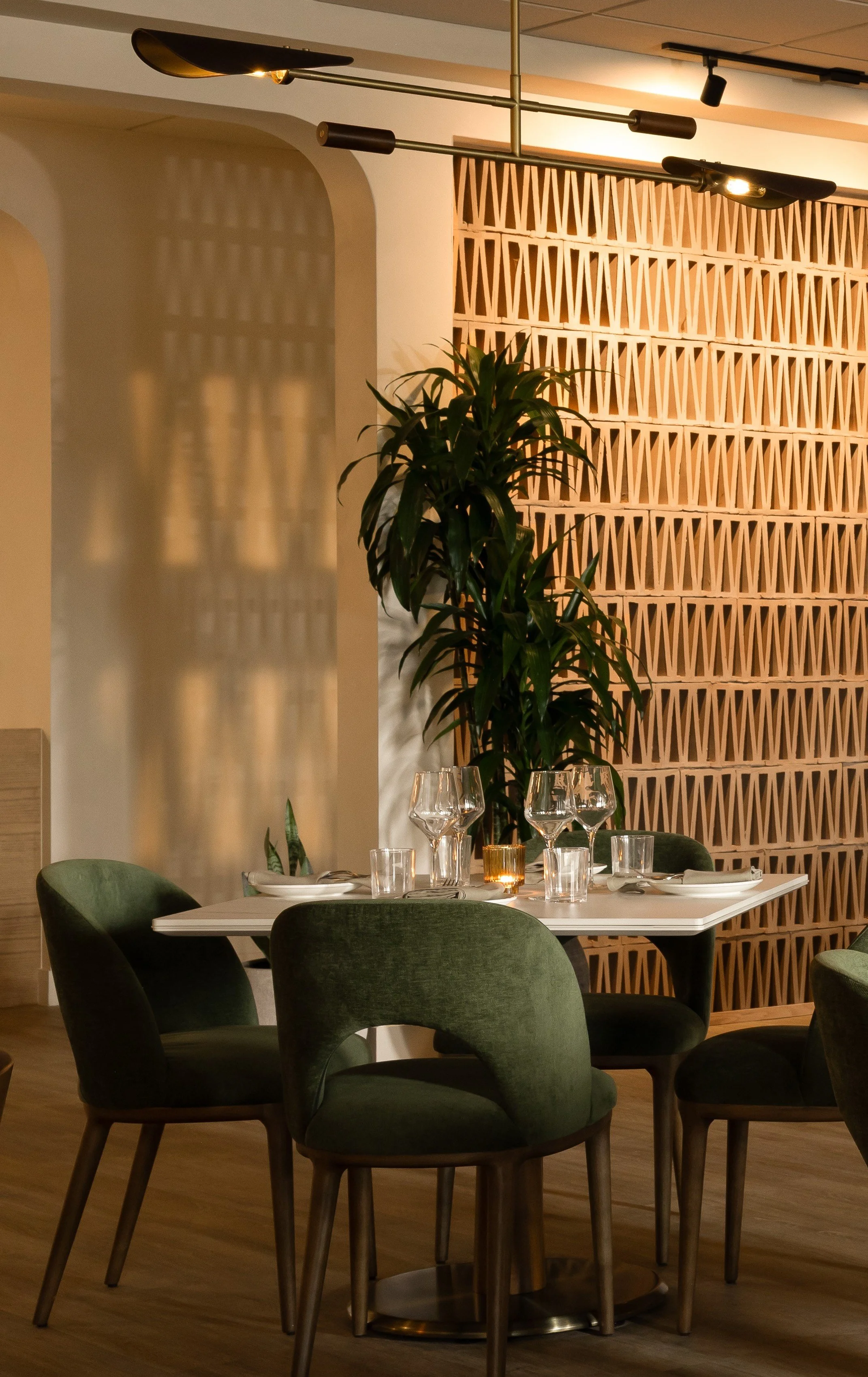 A table set for dining in a restaurant with green upholstered chairs, wine glasses, plates, and candles, featuring a wooden lattice wall and a tall leafy plant.