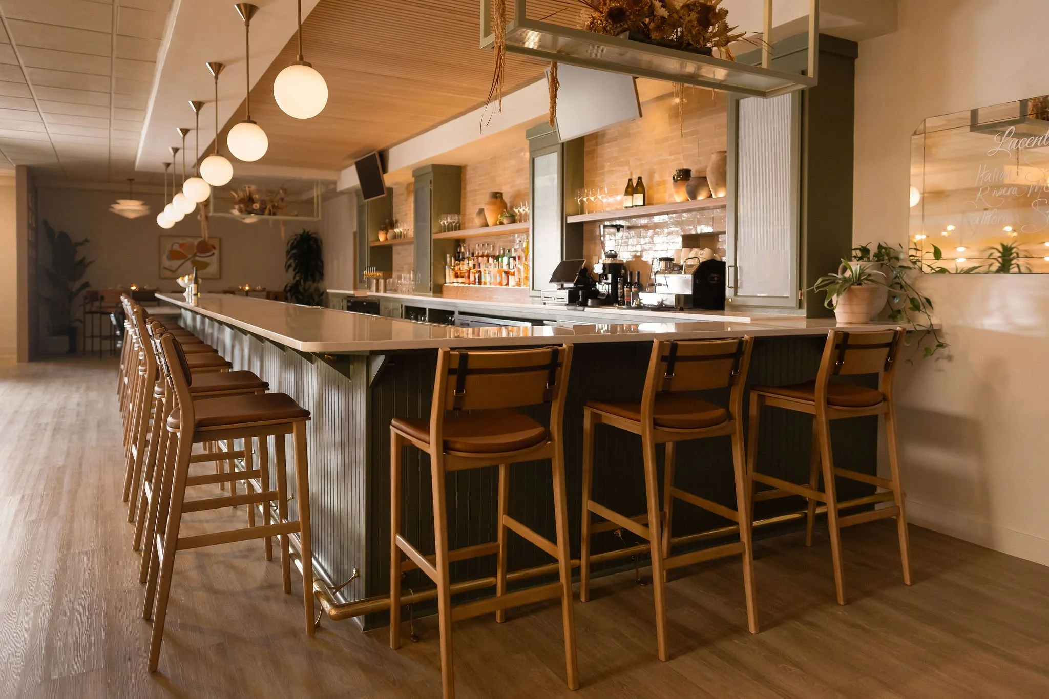A bar counter inside a restaurant with wooden bar stools, hanging globe pendant lights, shelves with liquor bottles and decor, coffee machines, and a glass display case. Plants and wall art are visible in the background.