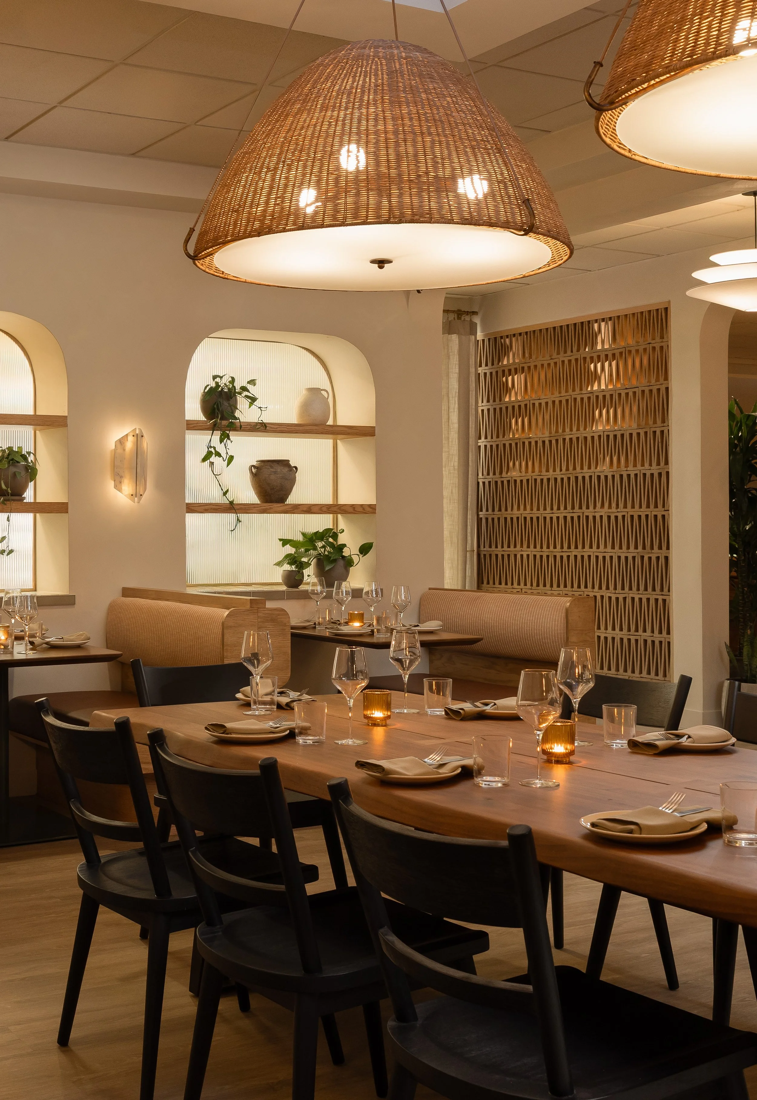A cozy restaurant interior decorated with warm lighting, hanging wicker lamps, plants, and wooden accents. The tables are set with glasses, plates, napkins, and candles, creating an inviting atmosphere.