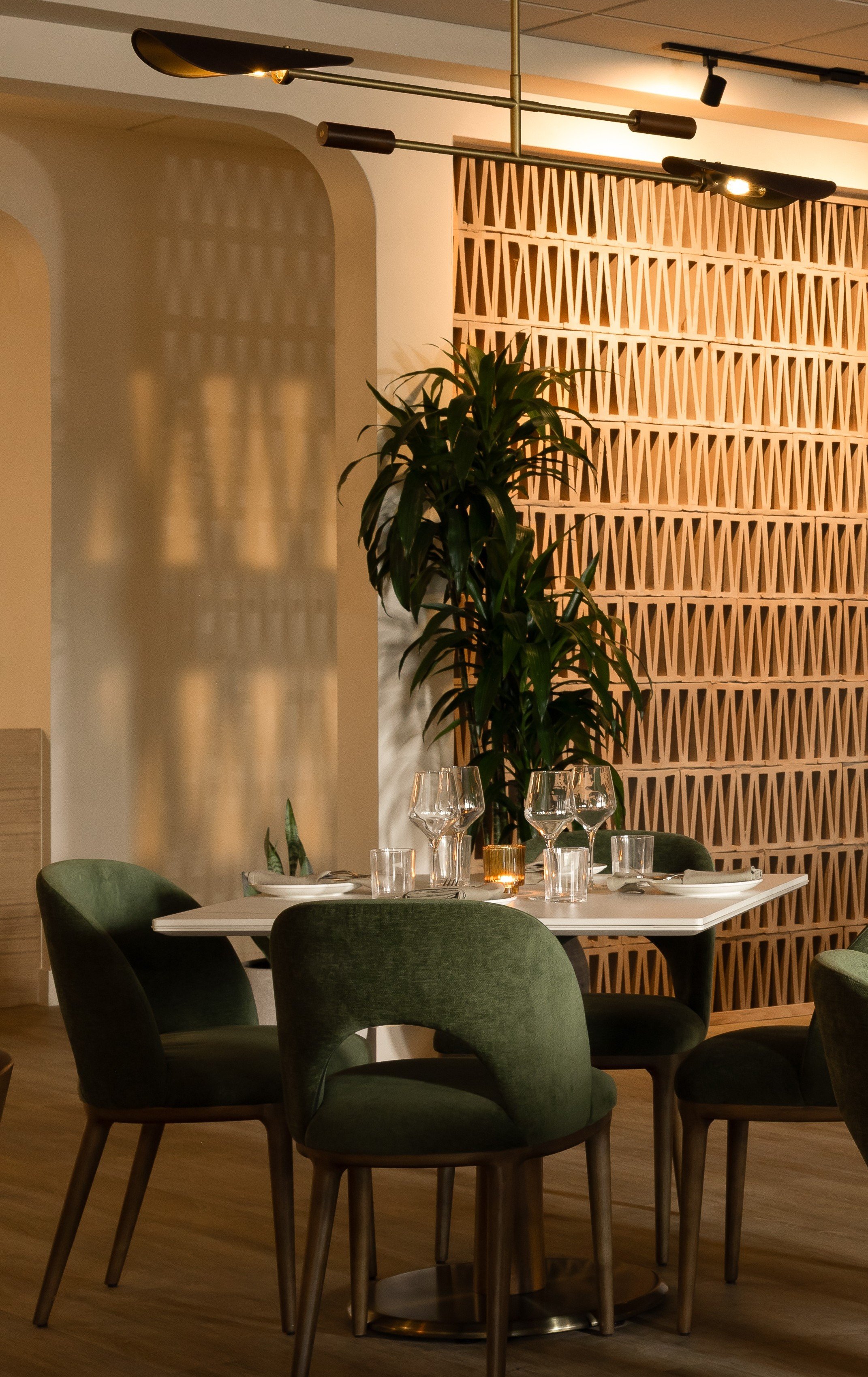 A modern restaurant dining area with a white table set for dinner, surrounded by dark green velvet chairs. There are wine glasses, candles, and tableware. A tall green plant and decorative wooden wall partition are in the background.
