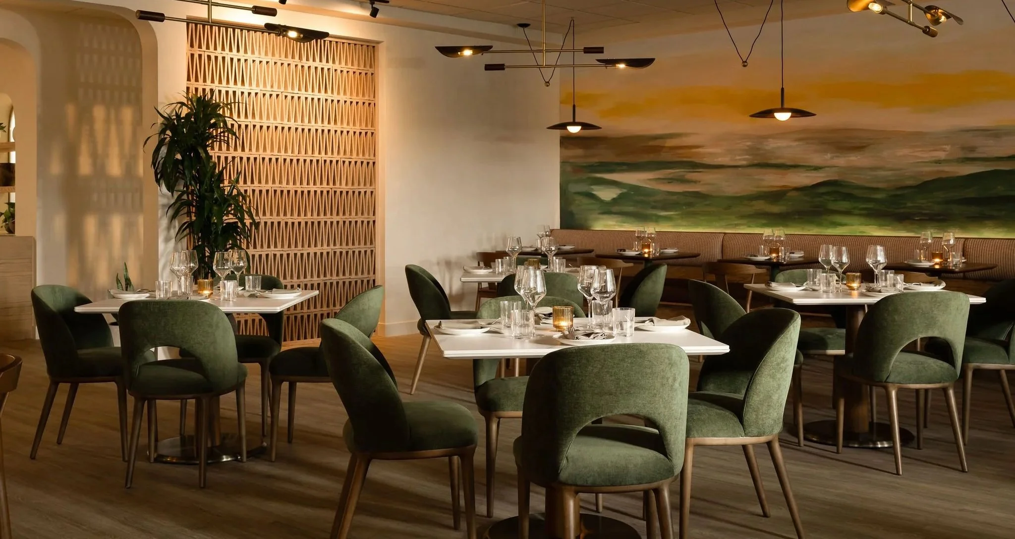 A modern restaurant dining area with green upholstered chairs, white tables set with glassware and napkins, candles, a large abstract landscape mural on the wall, and decorative ceiling lights.