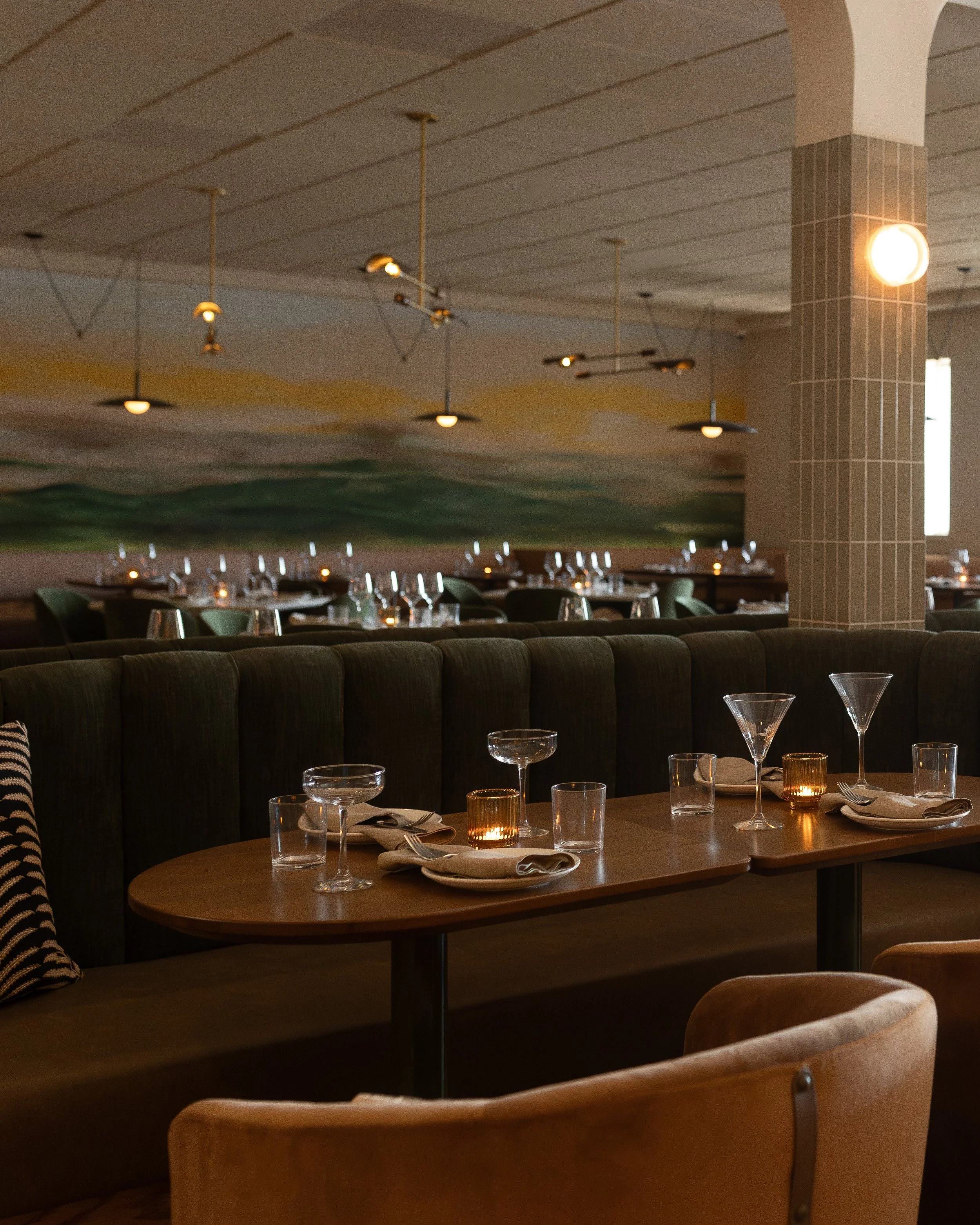 A restaurant or dining area with a wooden table set with glasses, napkins, and candles. Behind the table are green upholstered chairs and a cushioned beige armchair. The background features a mural with a landscape scene and modern ceiling lights. A 