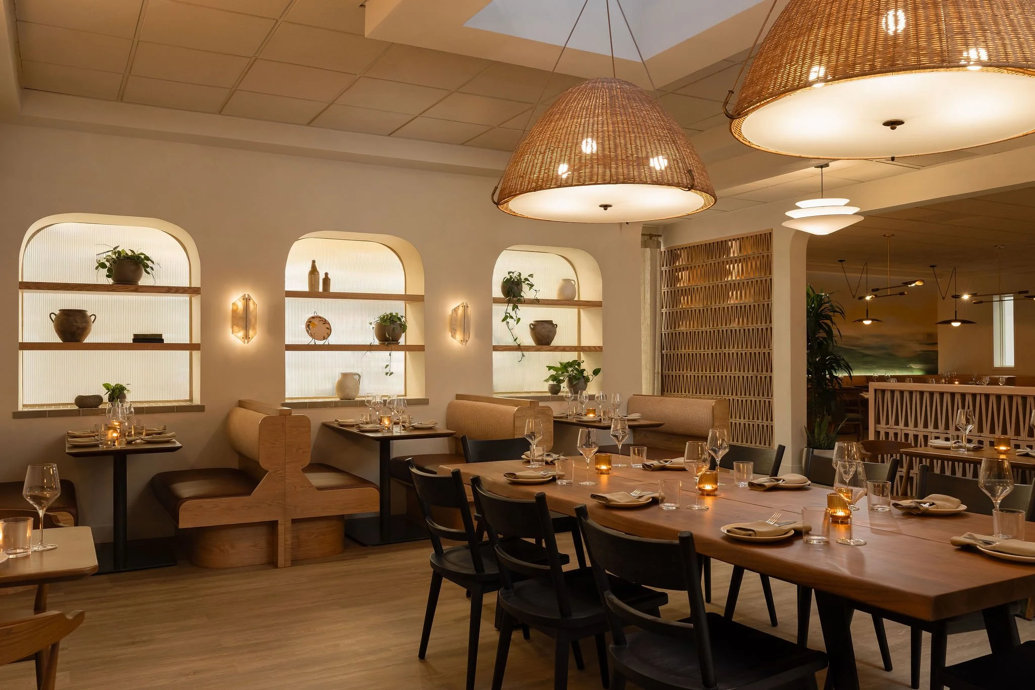 Interior of a restaurant with wooden tables set with glassware, napkins, and tealight candles, surrounded by black chairs and beige booth seating, decorated with potted plants, pottery, and wall-mounted light fixtures, illuminated by hanging woven an