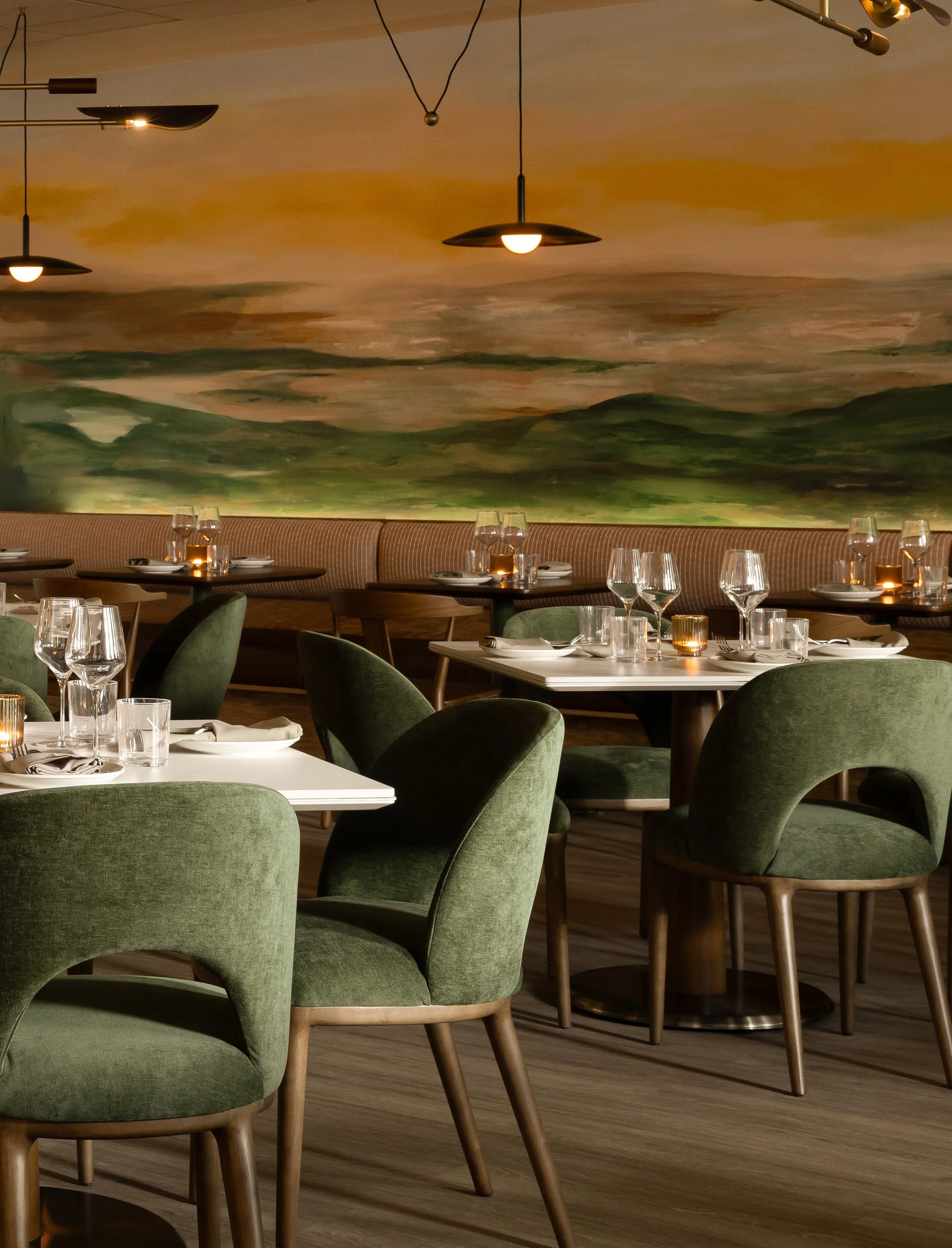 Interior of a restaurant with green upholstered chairs, tables set with white plates, napkins, and wine glasses, a colorful landscape mural on the wall, and modern ceiling lights.