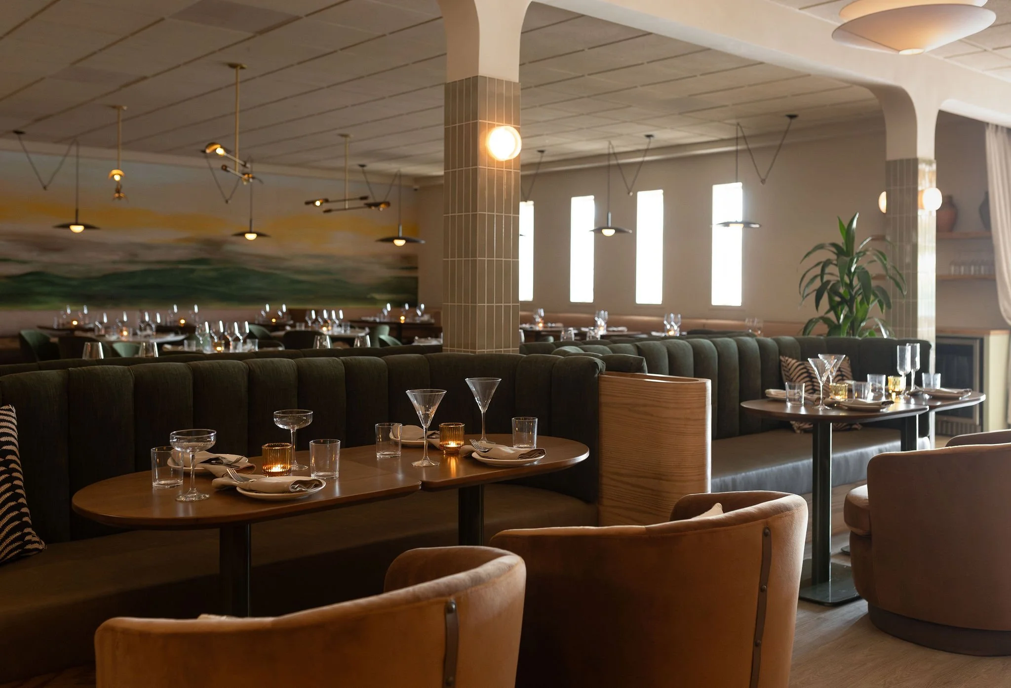 Interior view of a modern restaurant with tables set for dining, featuring wine glasses, candles, and napkins, with large windows and artistic ceiling lights.