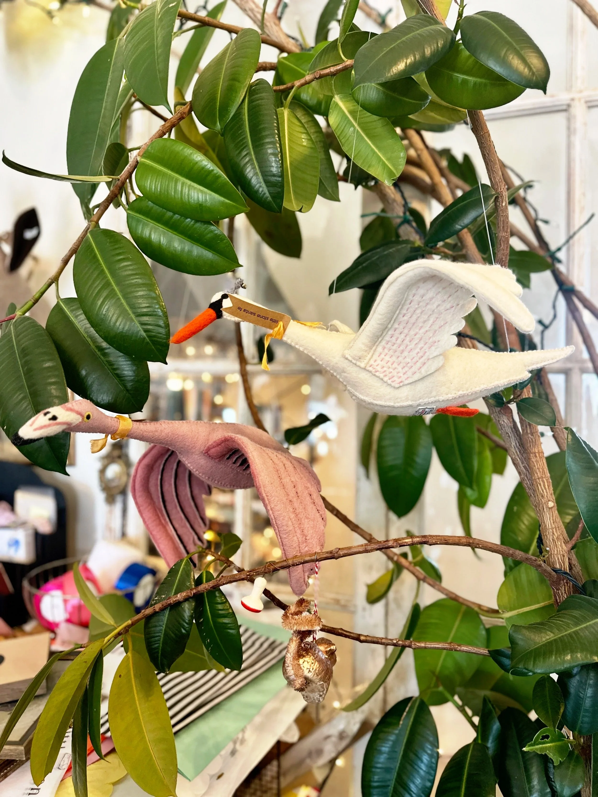 Fabric plush toys of a stork, a flamingo, and a squirrel hanging from a tree with green leaves inside a store.