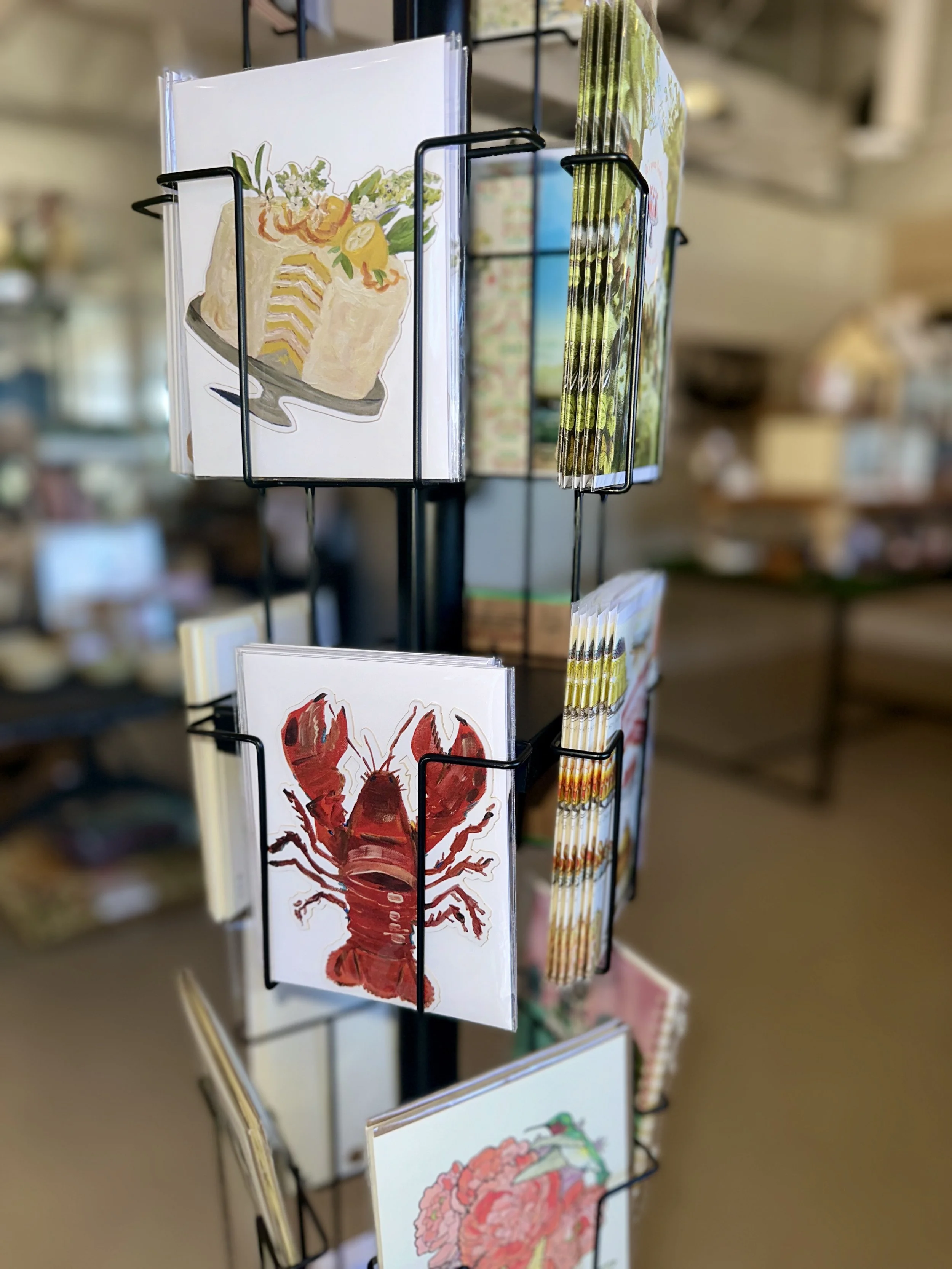 Postcards with illustrations of a cake, a lobster, and a flower, displayed on a black rotating rack in a store.