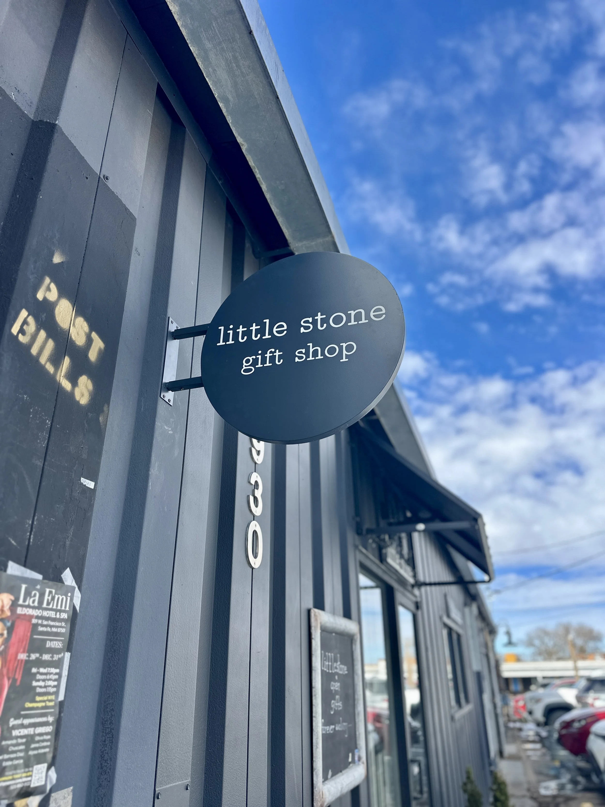 Black round sign that reads "little stone gift shop" in white lowercase letters, mounted on the exterior wall of a building under a blue sky with scattered clouds.