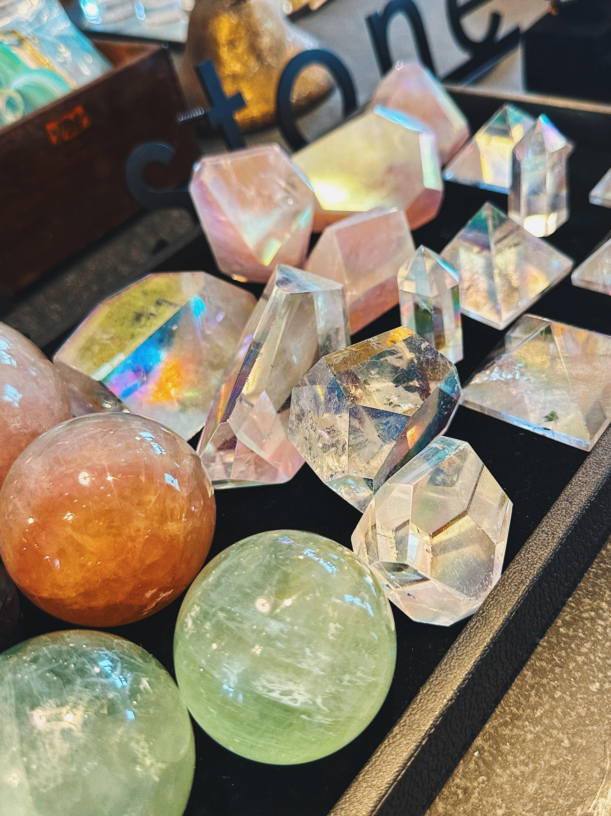 Display of various colorful, iridescent geometric crystals on a black surface, with a wooden sign and a gold-colored vase in the background.